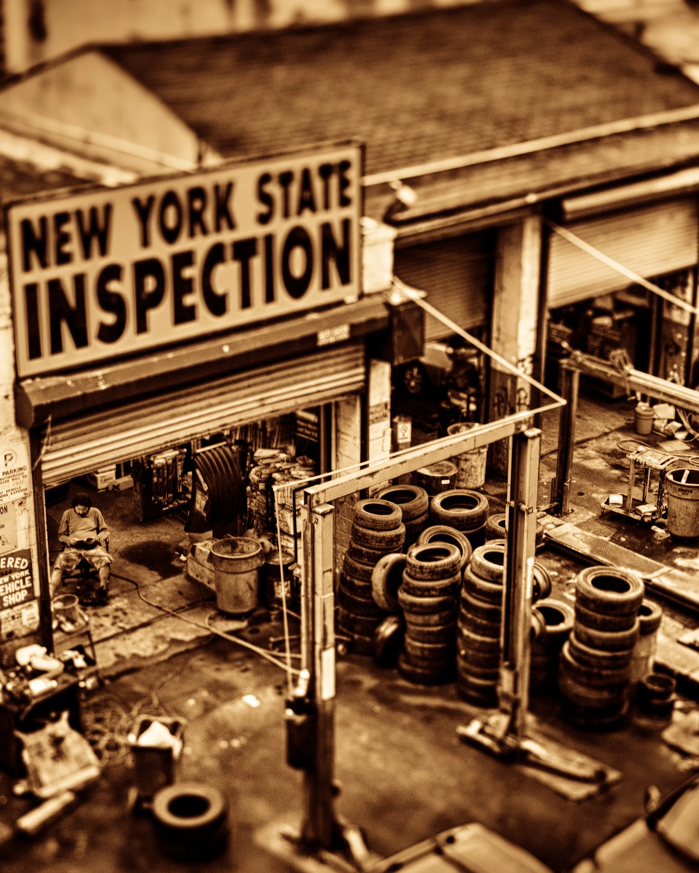 Sepia toned fine art photograph looking into a former New York State inspection and tire shop in Manhattan, with a man seated quietly inside the garage.