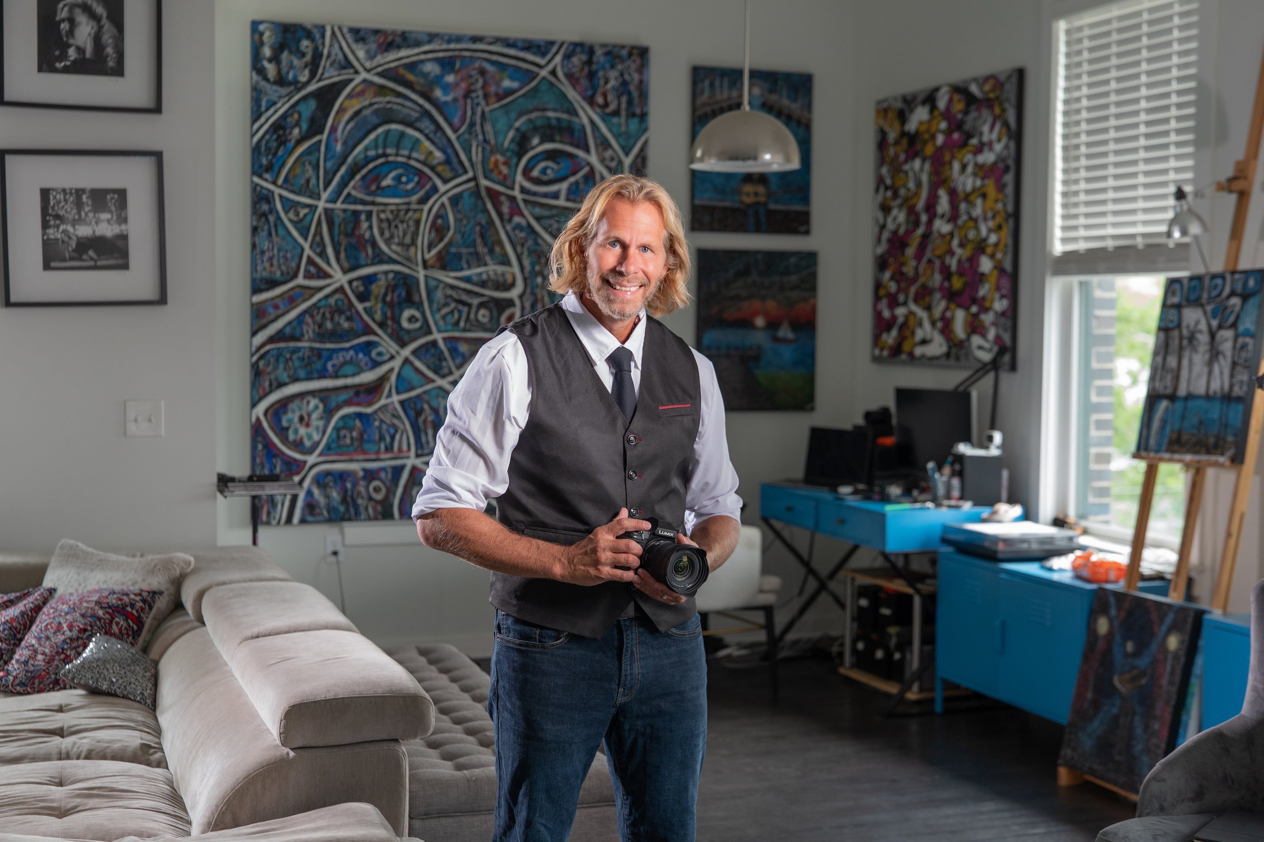 Charles Maring, keynote speaker, photographer, filmmaker, and visual artist, holding a camera in his Savannah art studio surrounded by original paintings.