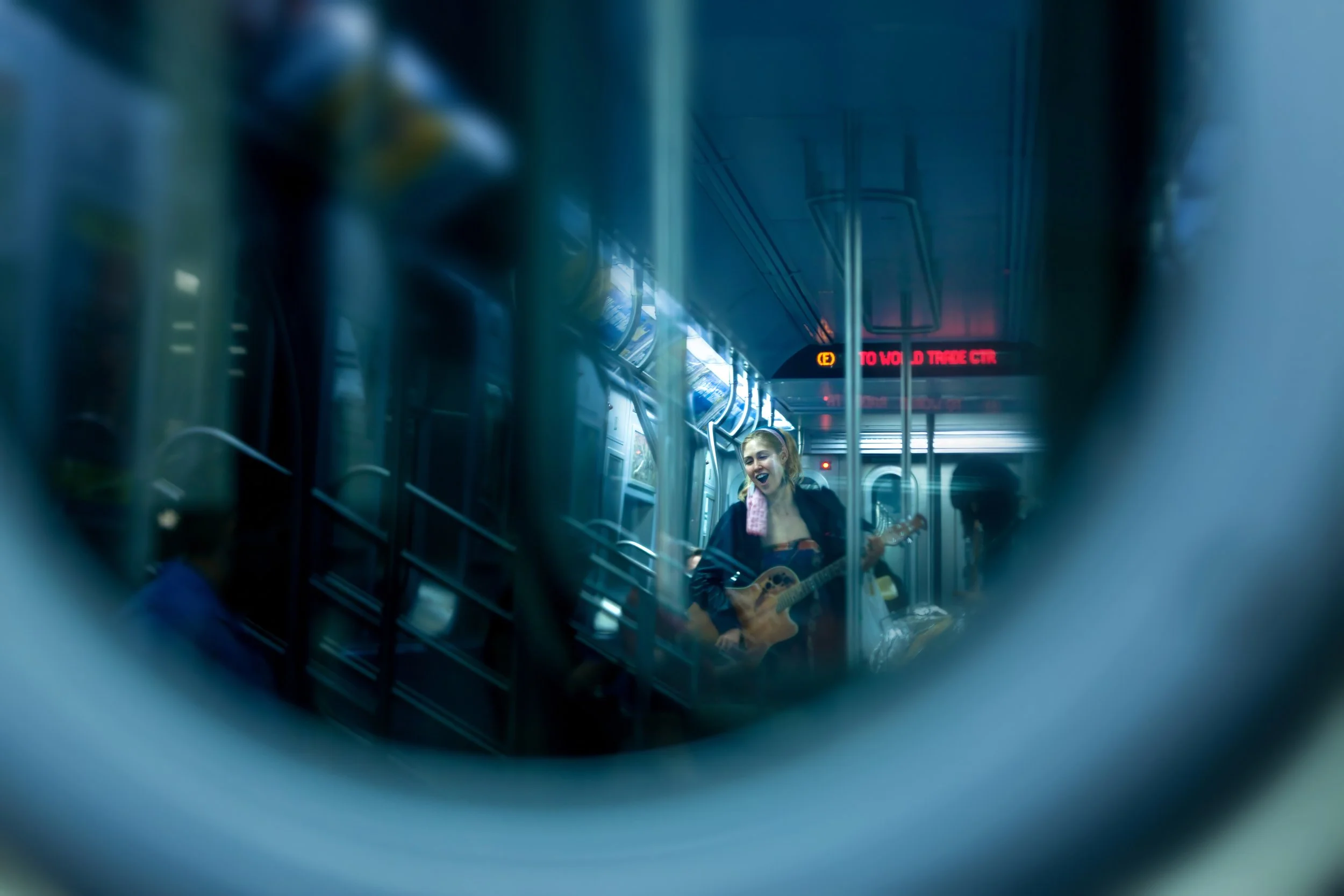 Fine art photograph of a woman playing acoustic guitar and singing in a New York City subway car, seen through the window of the adjoining train car.