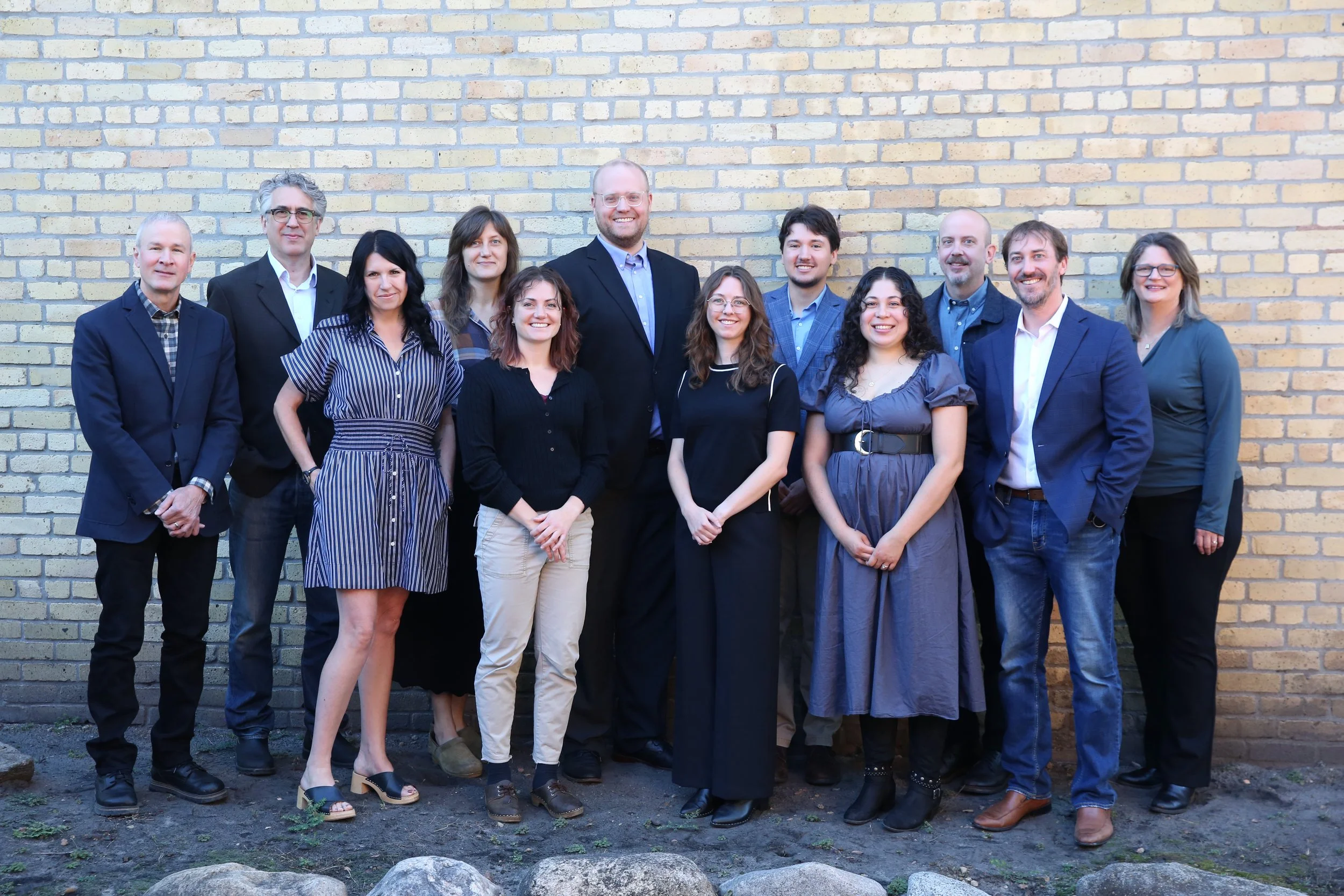 A diverse group of twelve people, both men and women, standing side by side against a beige brick wall outdoors. They are dressed in business casual attire, smiling, and appear to be posing for a group photo.