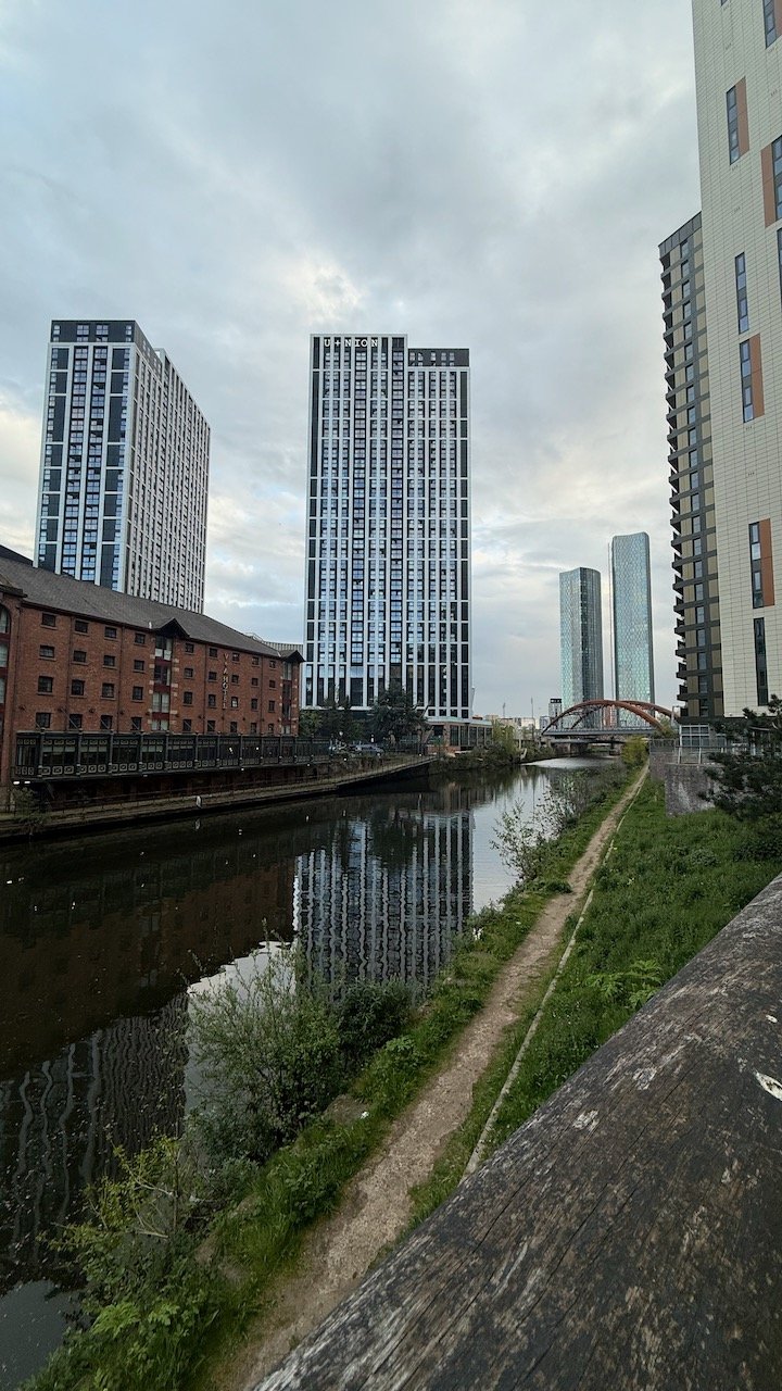 Canal-side view towards apartment towers in Manchester and Salford beneath a cloudy sky.