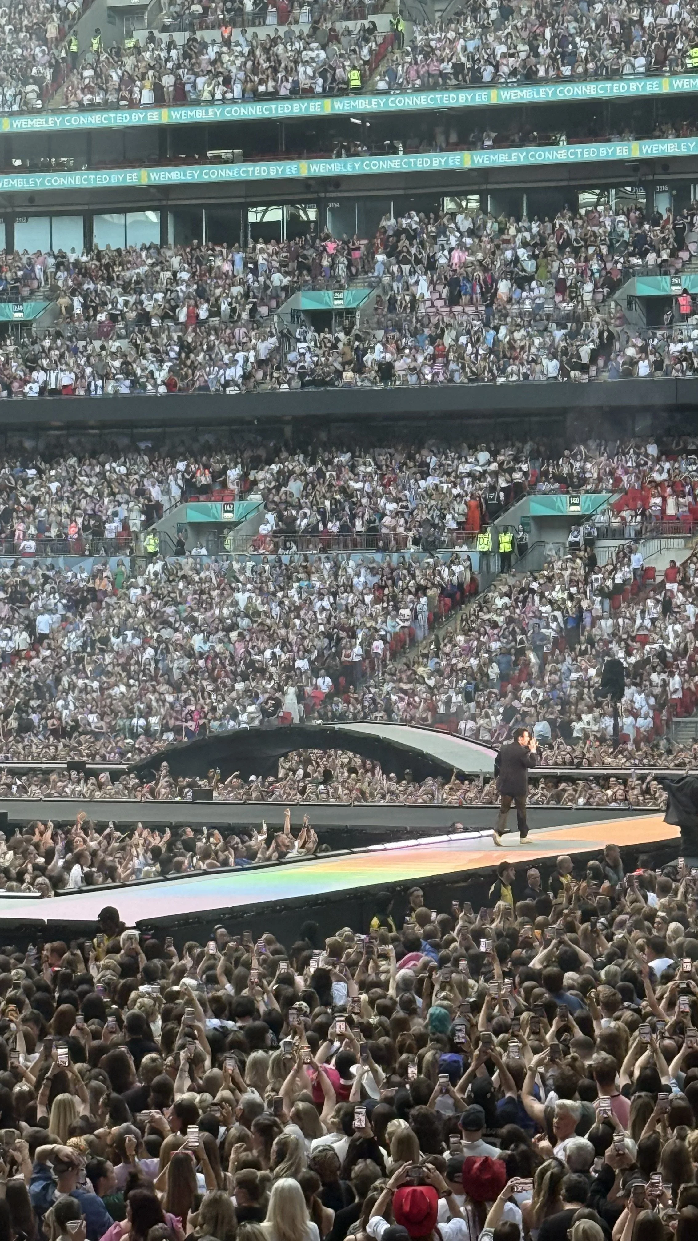 Harry Styles performing on the rainbow-lit Wembley Stadium catwalk, surrounded by a large crowd of fans holding up phones.