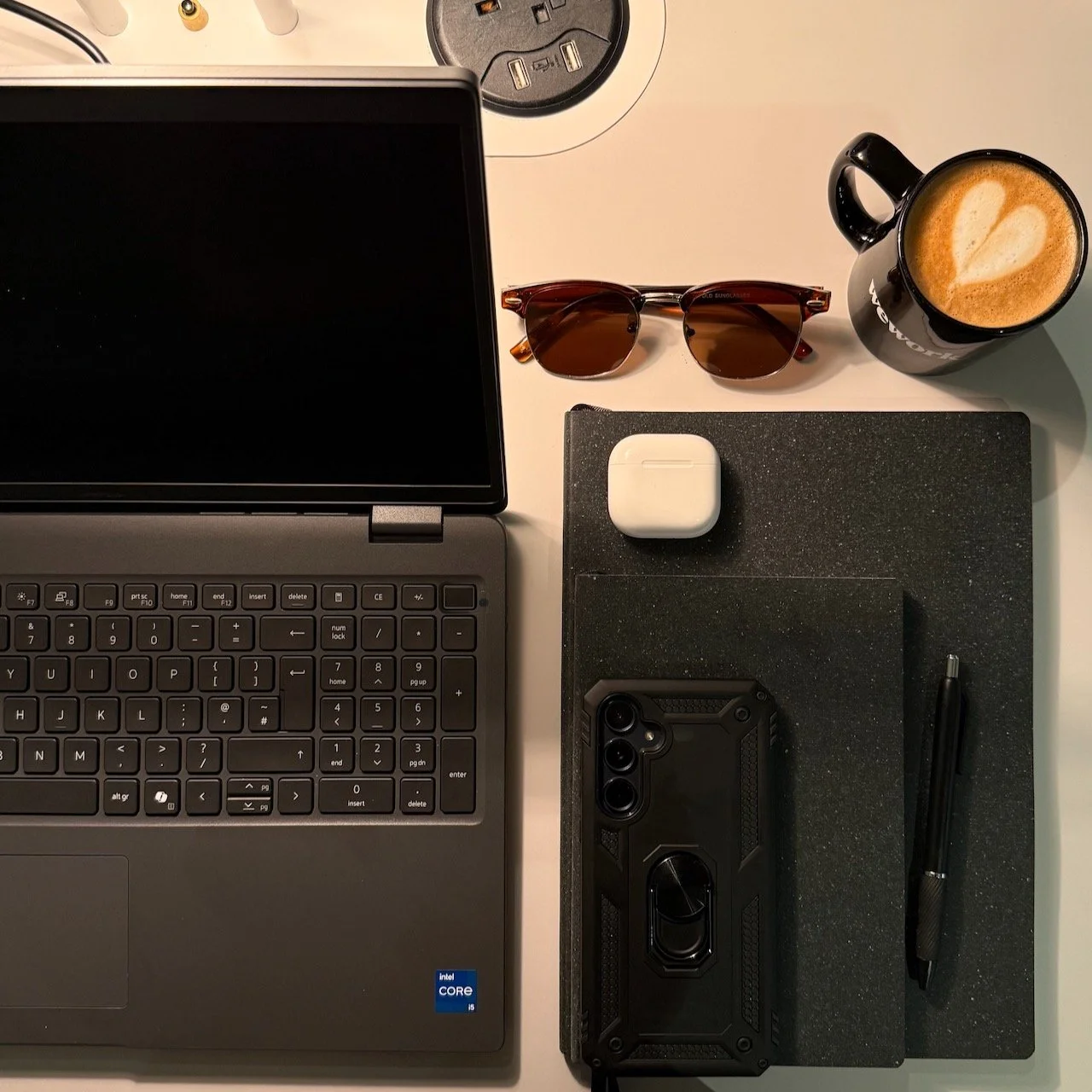 Laptop, notebook, phone, sunglasses and coffee on a desk during Greg Corrigan's first week in a UK communications role.