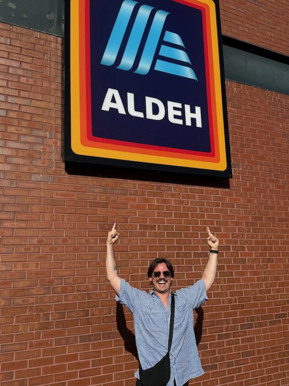 Greg Corrigan smiling and pointing at an 'ALDEH' sign before the Oasis concert at Heaton Park.