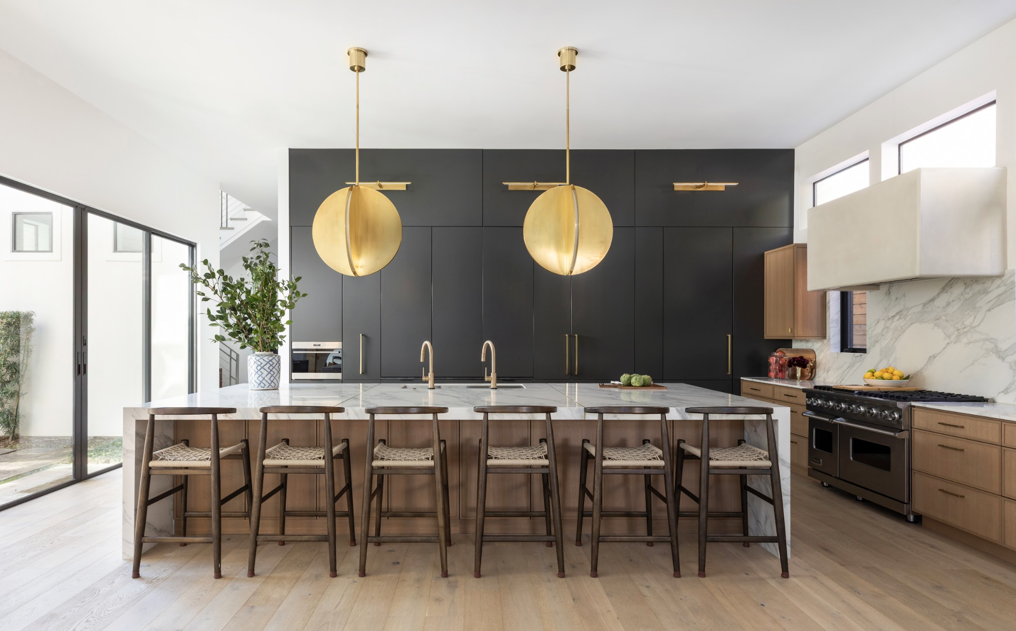 Modern kitchen with black cabinets, white marble island with wooden bar stools, two large gold pendant lights, and light wood flooring.