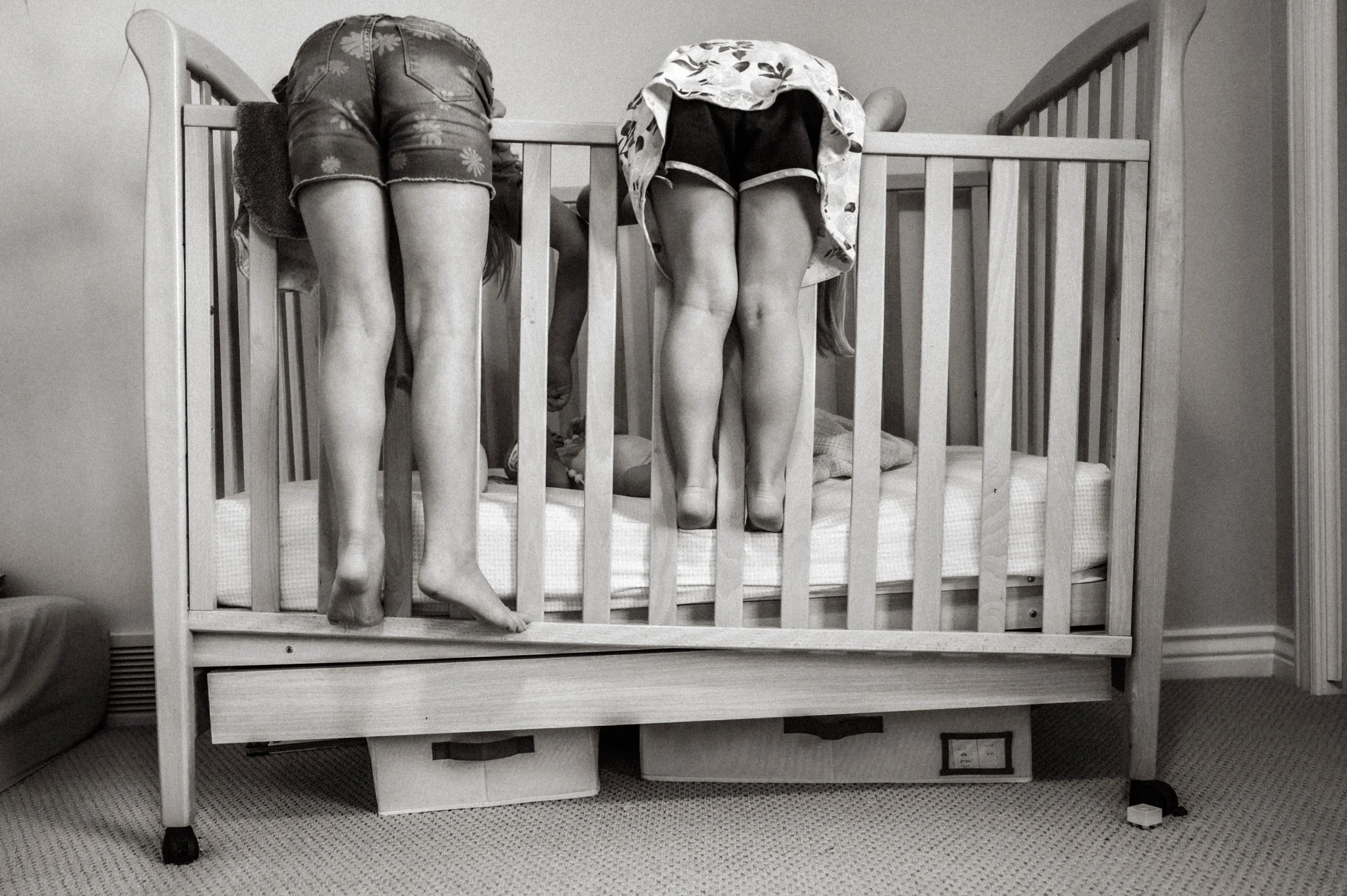 A candid moment of two young sisters leaning over a baby's crib during a family story session.