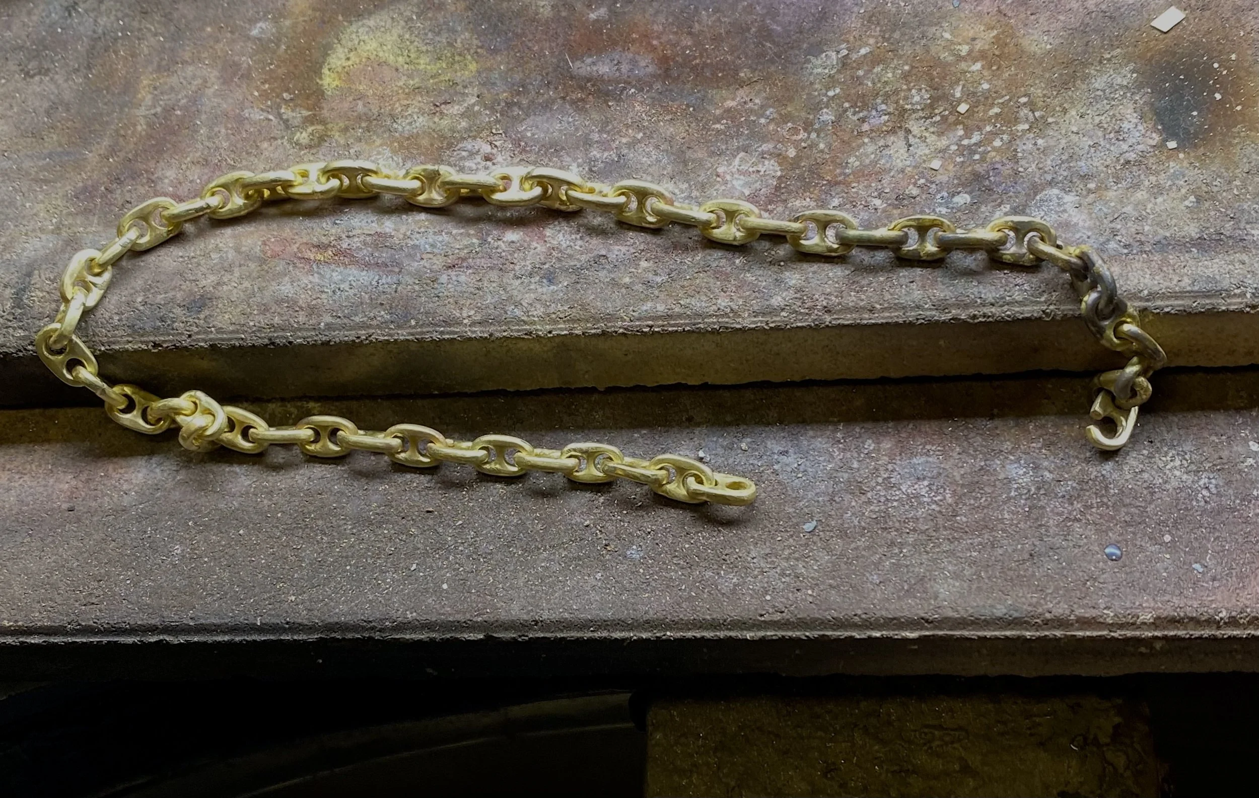 Close-up of a gold nautical chain laid on a rough, textured surface in a workshop.