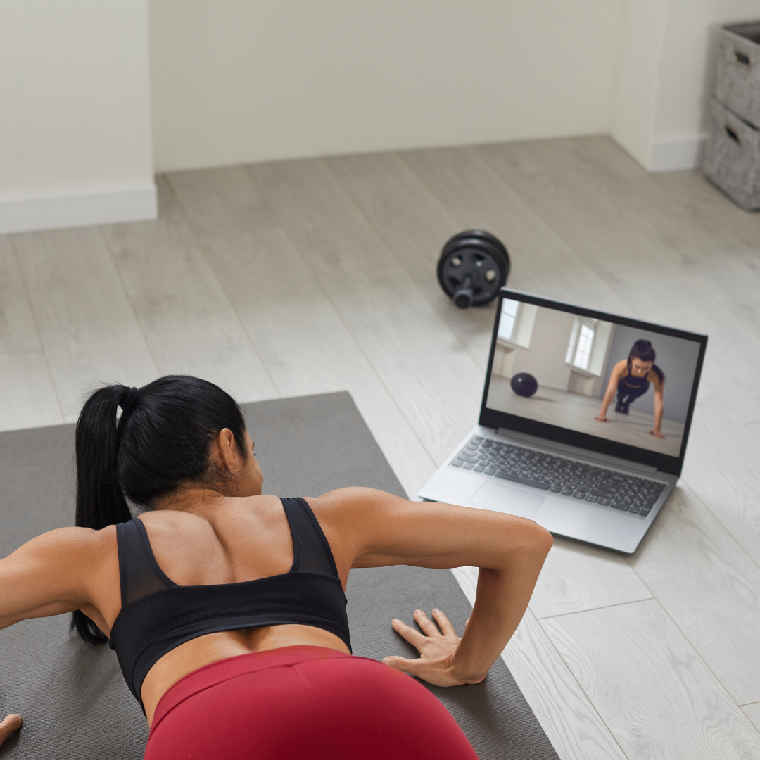 Woman doing push-ups on a yoga mat in front of a laptop showing a workout video, with an exercise wheel nearby.