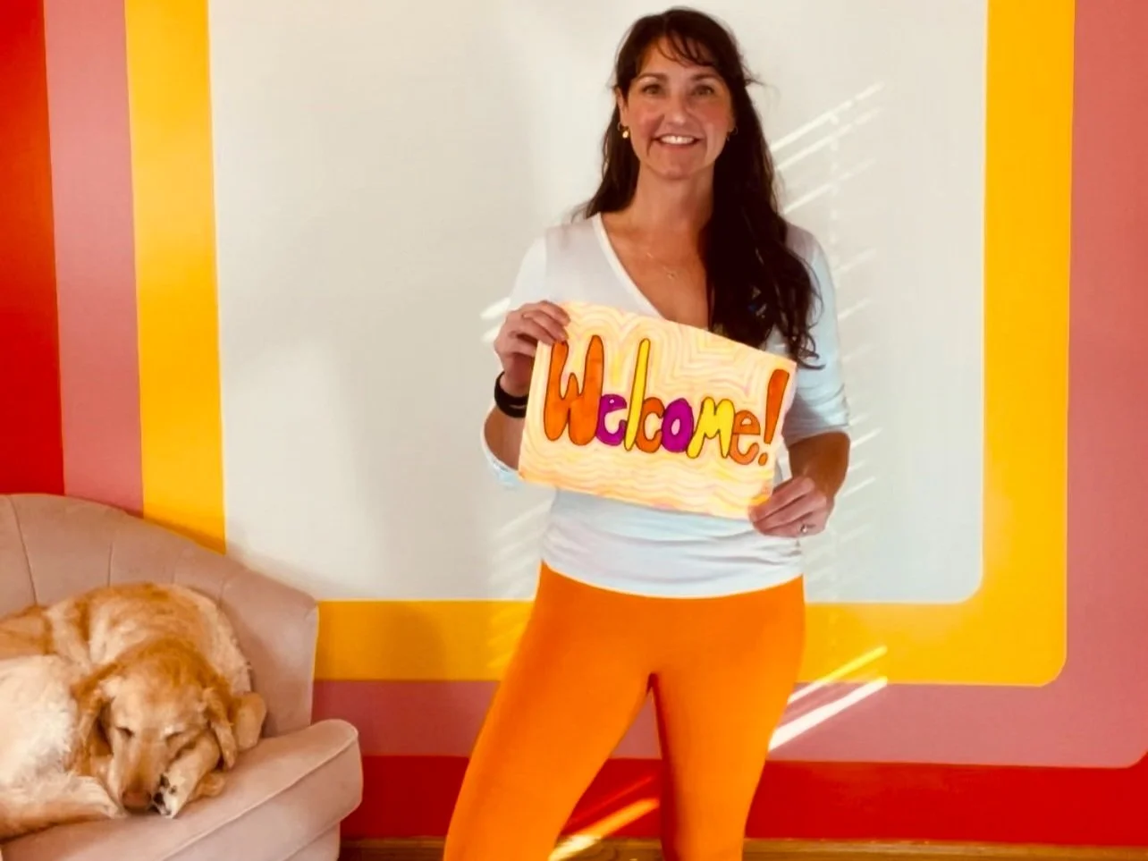 A woman stands indoors holding a colorful "Welcome" sign. She is wearing a white top and orange pants. Next to her, a golden retriever dog is sleeping on a beige chair. The wall behind has red, yellow, and pink stripes.