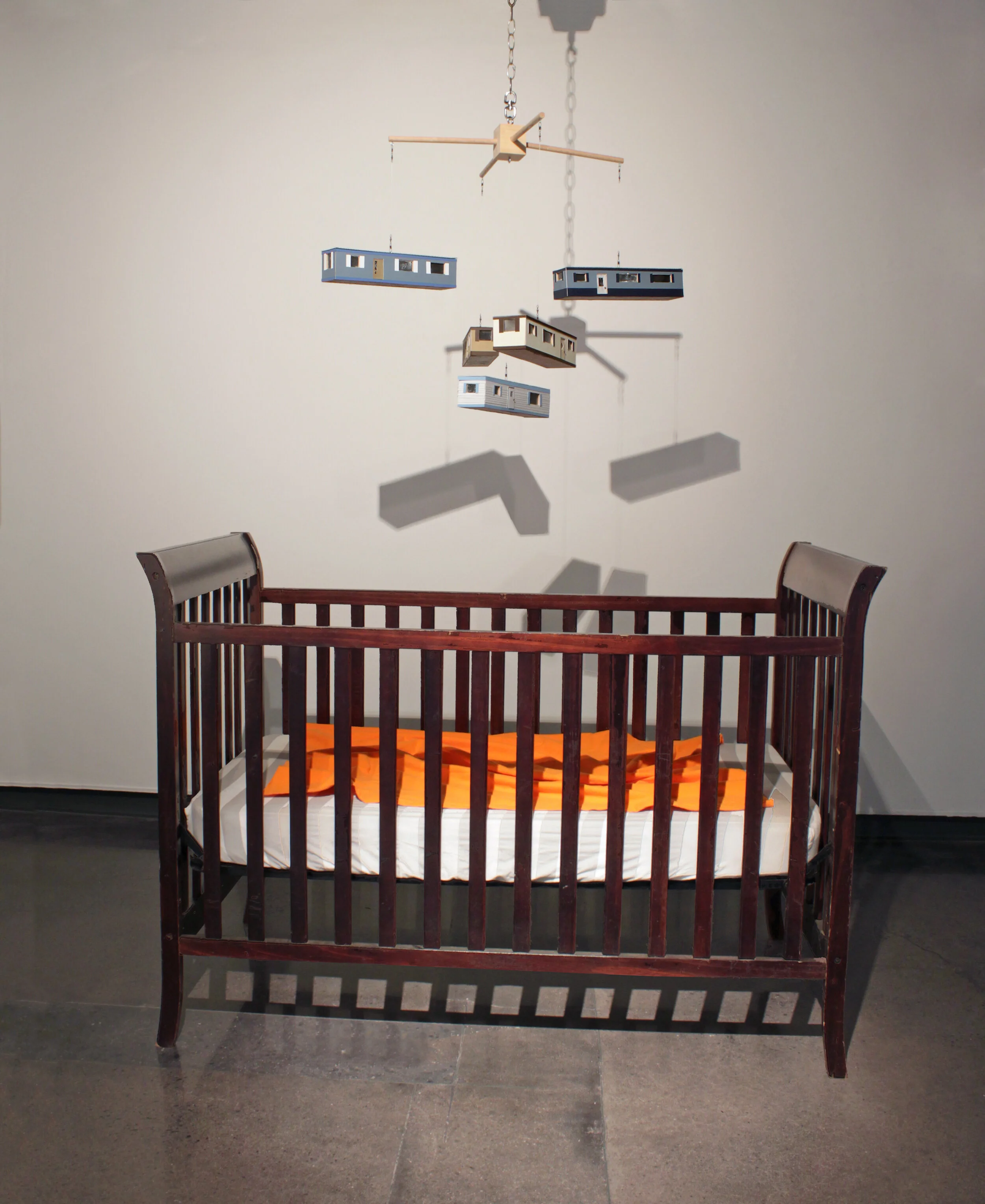 Confined. 2021. Found objects, mixed media. Confined reflects the metaphorical cage in which children can become trapped when growing up in a disadvantaged socioeconomic situation. This visual representation is a mobile-home-mobile that hangs above a baby crib. I felt that the mobile needed the added association of the doom of being raised in poverty, so the crib not only serves as an association for childhood but also represents a “cage” of destiny for most raised in this environment: a brutal cycle of the lower class.&nbsp;The crib has sheets with grey and white stripes to evoke old-time prison uniforms. The stripes also mimic and reinforce the jail-like structure of the barred crib. Included in the crib is a bright orange baby blanket that references the jumpsuits of modern-day prison inmates. The mobile-home-mobile is attached to a slow rotating motor. The circular, cyclical cycle of the mobile reinforces the idea that poverty can be a vicious cycle that persists over several generations of a family.