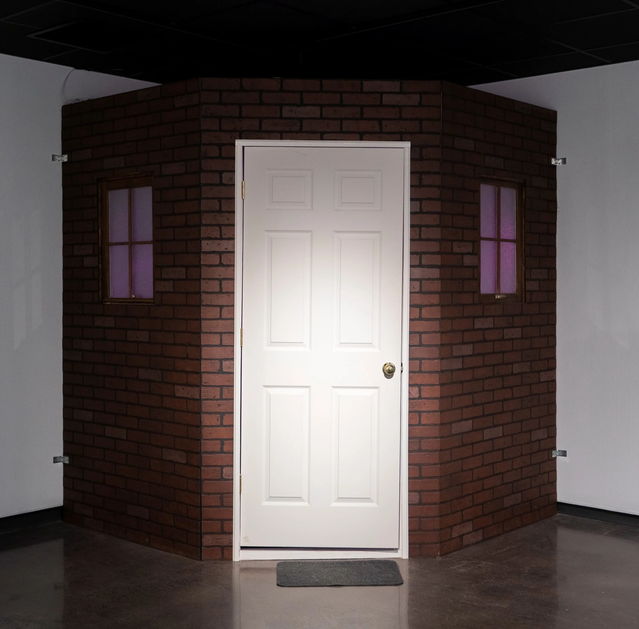 Housed. 2021. Wood, mirror, mixed media. Housed is an installation that explores the complex dynamics of trauma. It is a literal façade of a home on the exterior and an interactive light experience on the interior. The interior space is activated when someone opens the door and explores the inside. The exterior is faux brick and alludes to a safe habitat, implying a sturdy, immaculate, and durable home life. To add to the curiosity, the interior contains another house structure suspended from the ceiling. The suspended house is part home and part gut-like cube that illuminates the entire space. On the interior walls are a panoramic mosaic of shattered mirrors to intensify the lighting and disfigure the viewer’s reflection.