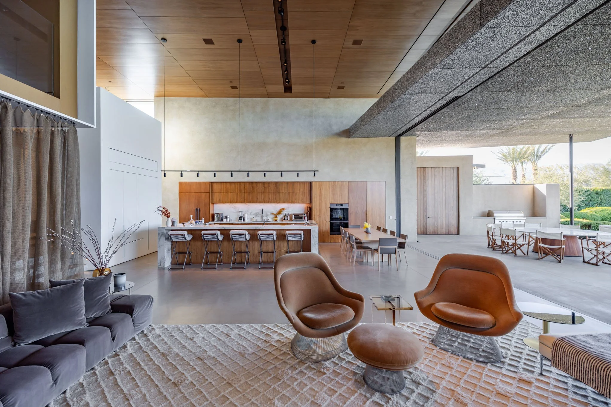 Open-concept living room and kitchen with modern furniture, wood and concrete finishes, and large windows showing outdoor seating and palm trees.