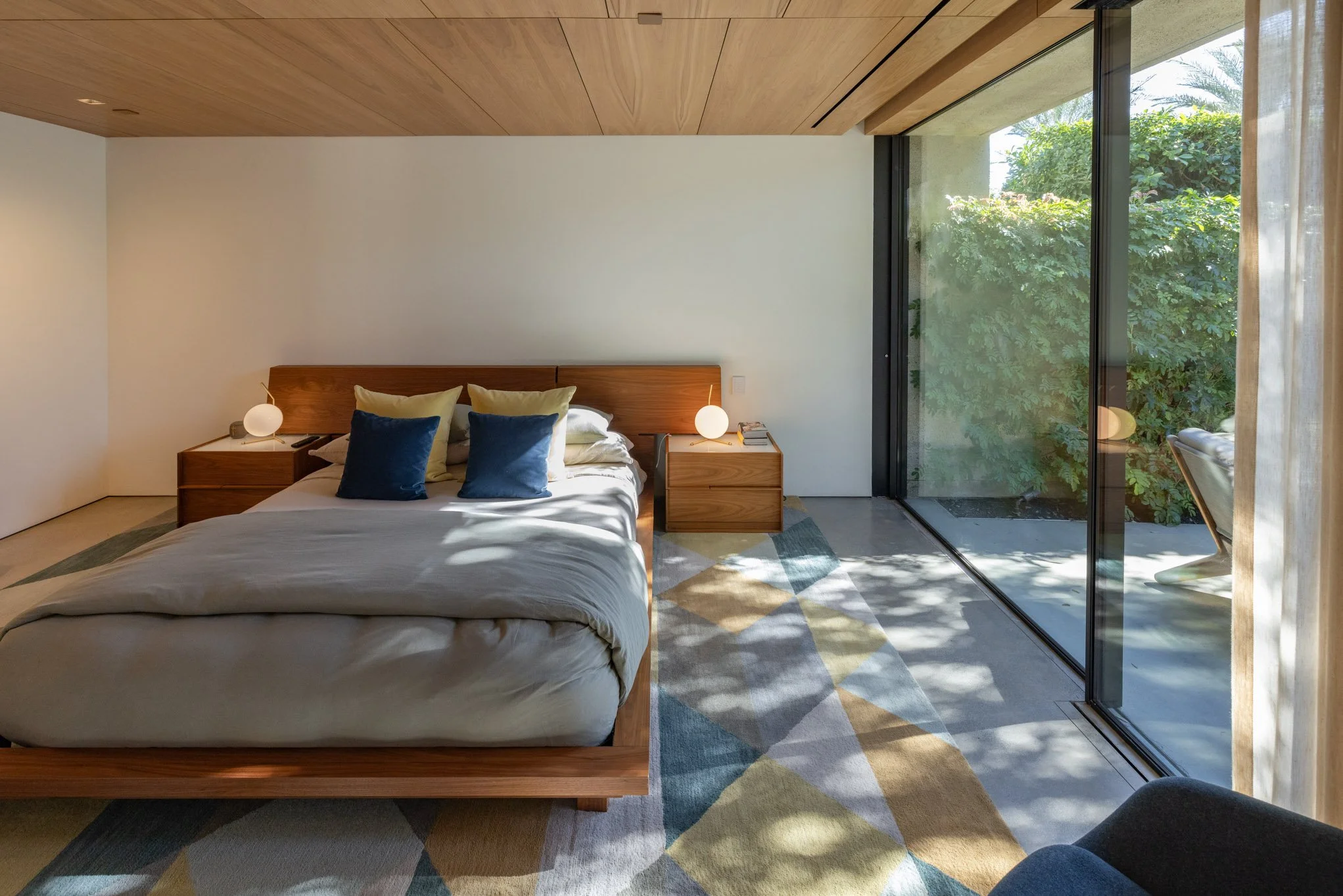 Modern bedroom with a large bed, two nightstands with lamps, a colorful geometric rug, and a large glass sliding door revealing greenery outside.