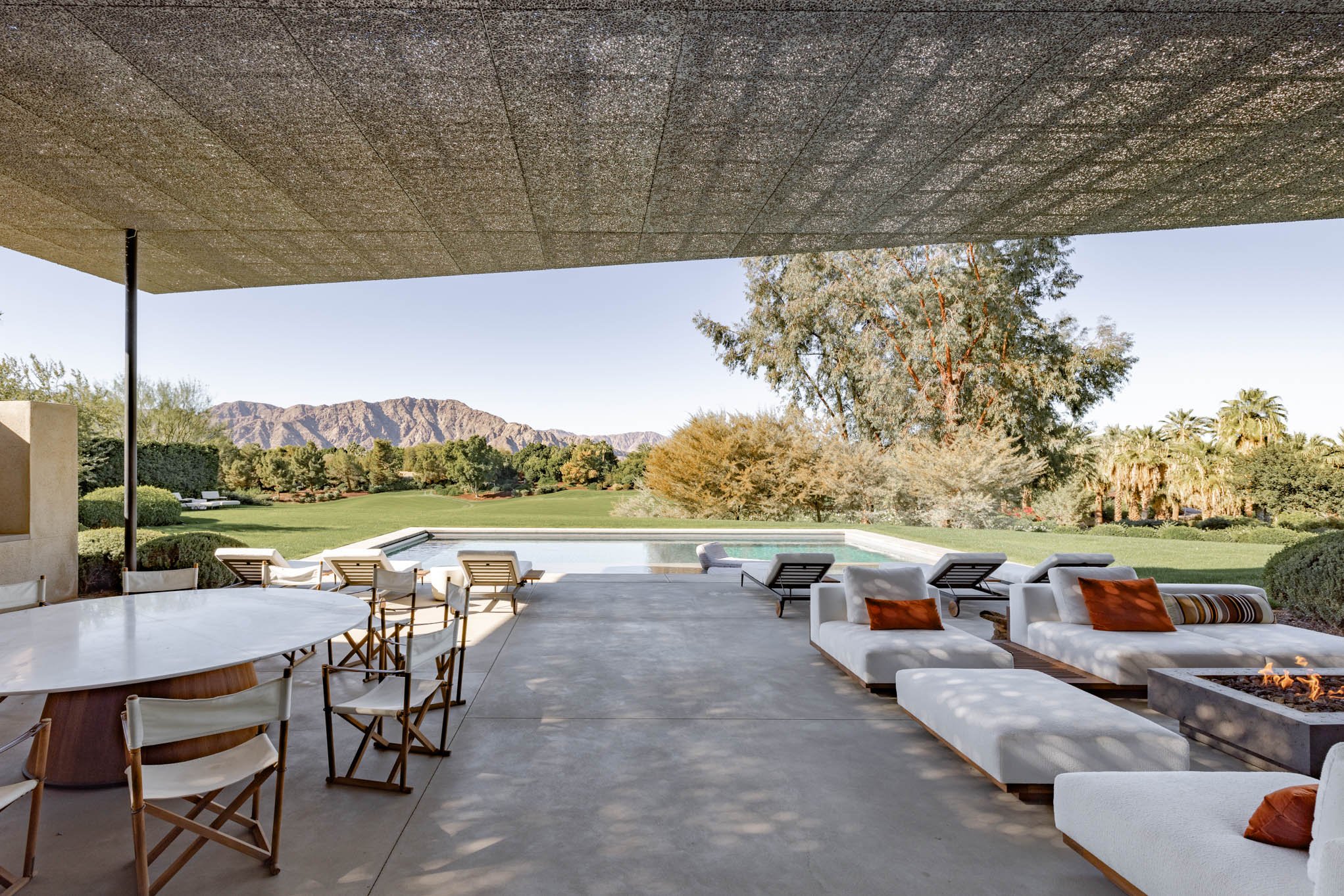 Luxury outdoor patio with lounge chairs, table, and fire pit overlooking a grassy area, trees, and mountains, under a covered ceiling.