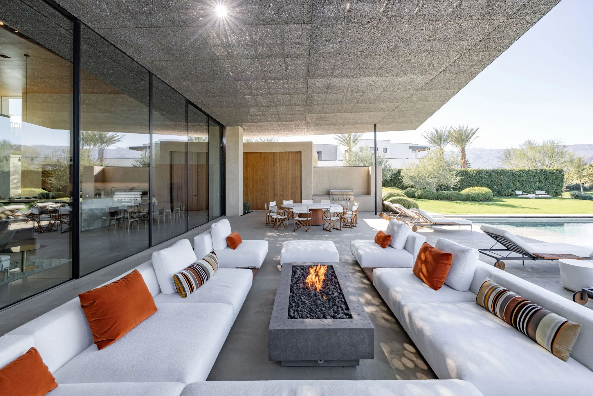 Outdoor modern patio with white couches, orange and striped pillows, a rectangular fire pit, black glass wall, and pool in the background, surrounded by greenery and palm trees.