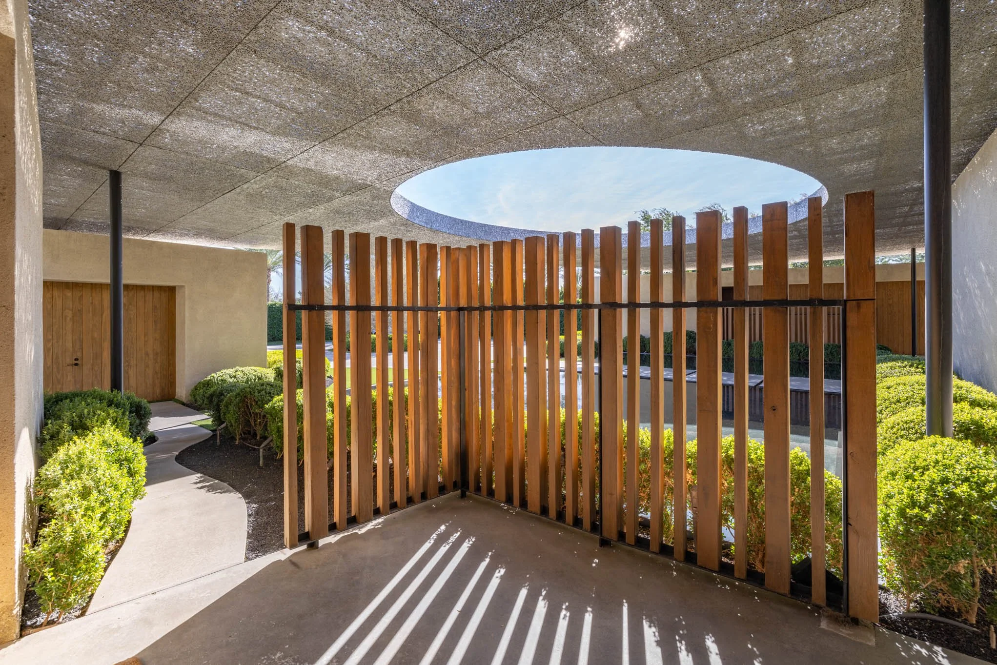 Architectural view of a modern outdoor space with a wooden slat fence, a concrete floor, and a curved concrete ceiling with a circular opening allowing sunlight. Green bushes and a pathway are visible.