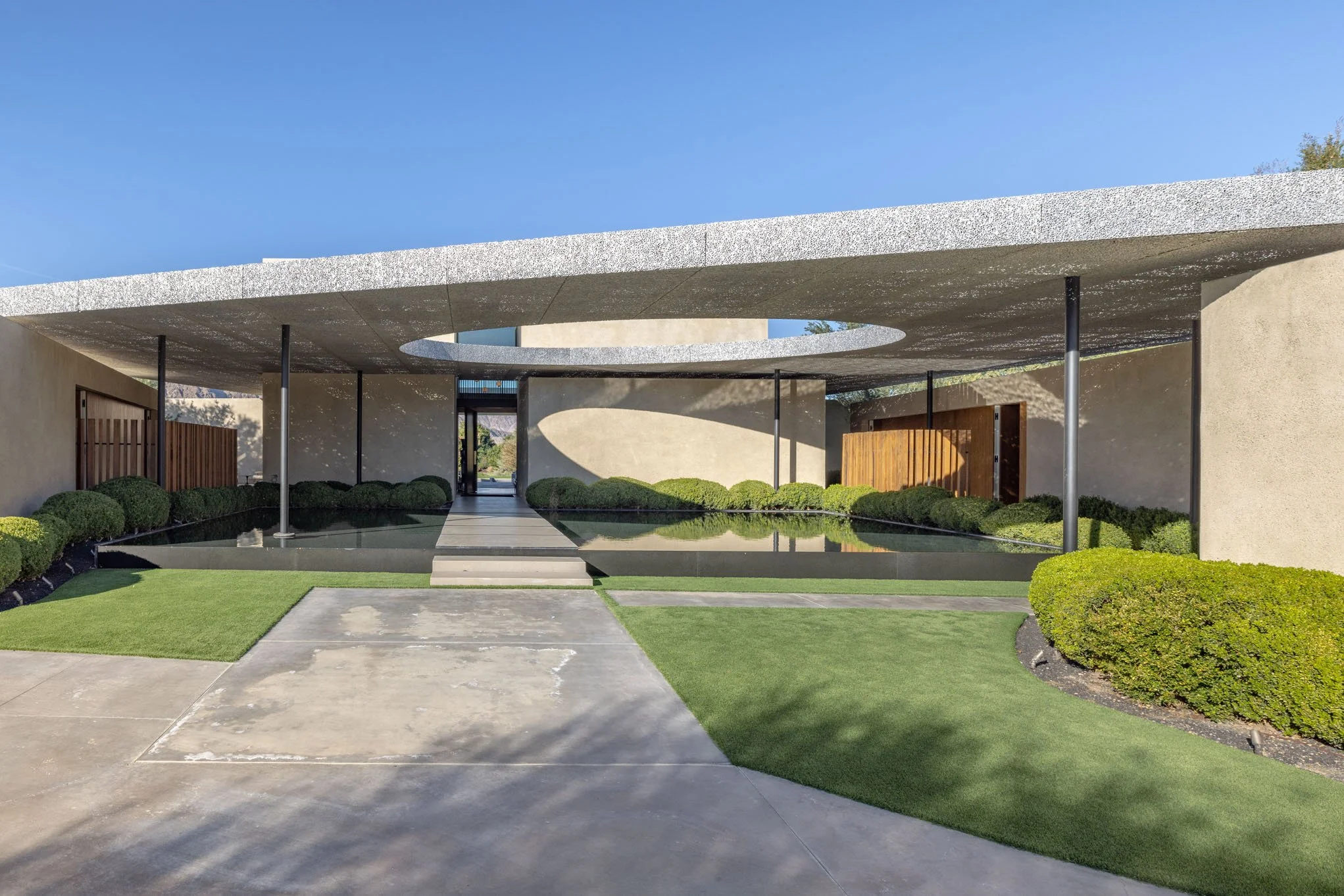 Modern architectural house with a concrete roof that has a circular opening, reflecting pool, green bushes, and a landscaped yard under a clear blue sky.