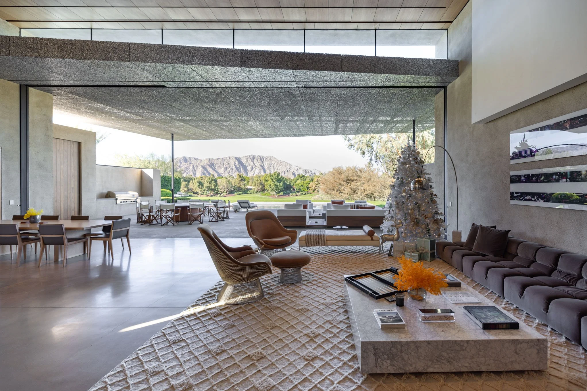 Modern living room with large open space, beige walls, and floor-to-ceiling windows overlooking a mountain landscape. Features a white Christmas tree, gray sofa, and various chairs, with a coffee table and media artwork on the wall.