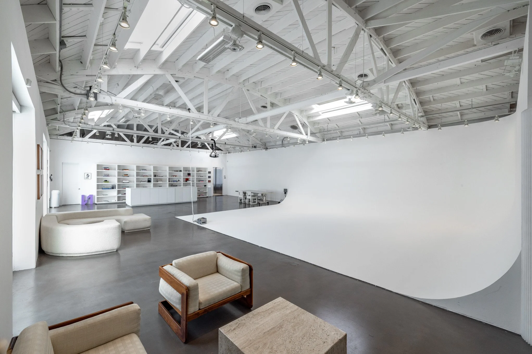 An empty photography studio with a white cyc wall, living room furniture, and a storage bookshelf in a high-ceiling space with exposed beams and skylights.