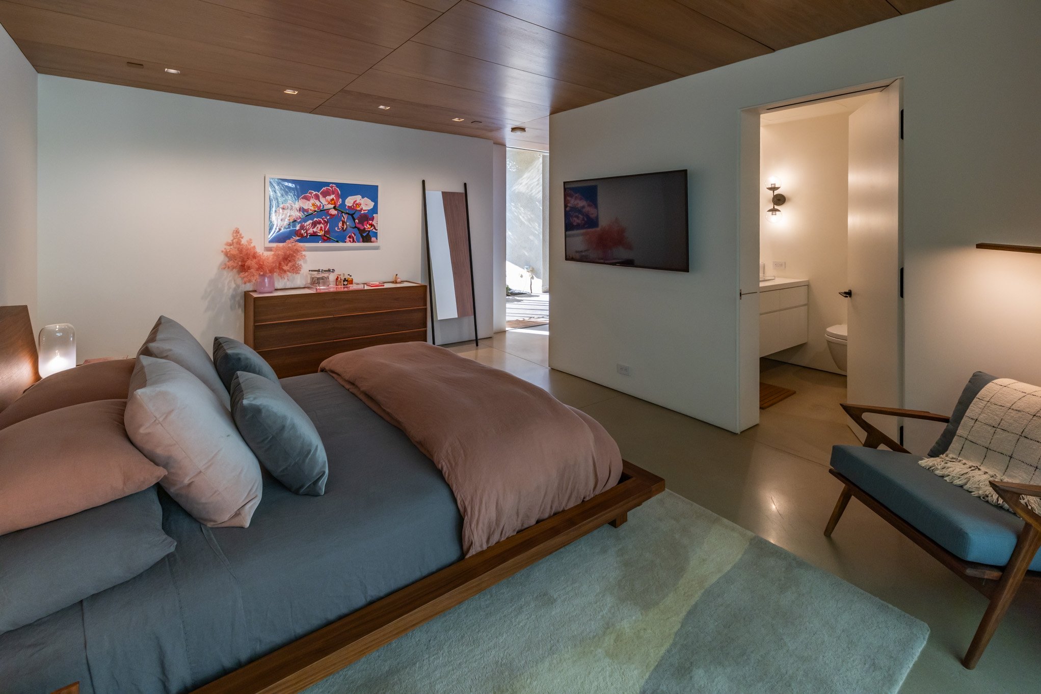 A modern bedroom with a neatly made bed featuring multiple pillows, a wooden dresser with pink flowers and artwork above, adjacent to a small sitting area with a chair and blanket, and a visible connected bathroom with lighting.
