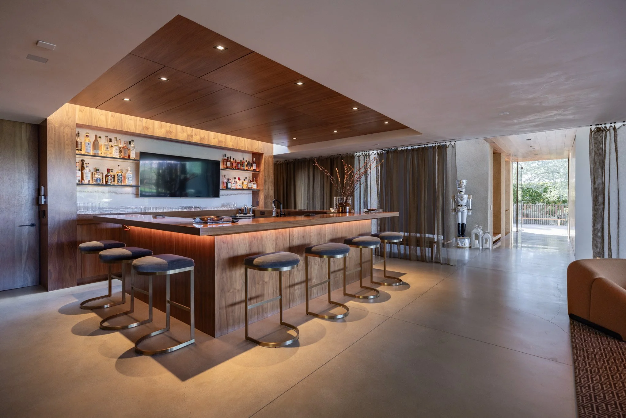 Modern home bar with a wooden countertop, bar stools, a TV, and shelves with liquor bottles, in a well-lit room with curtains and an outdoor view.