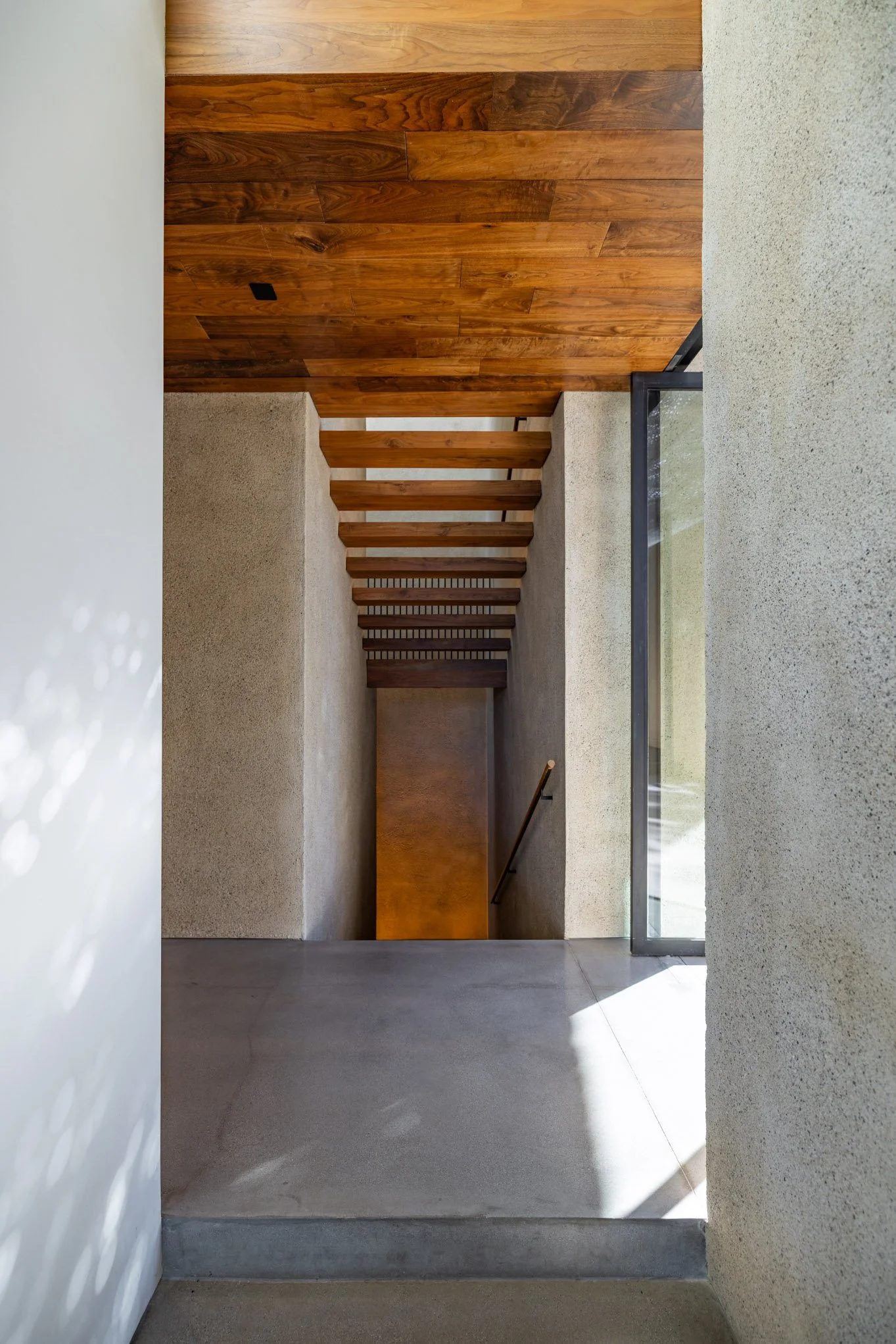 Interior view of a modern staircase with wooden steps, concrete walls, and a large glass window on the side, leading downward.