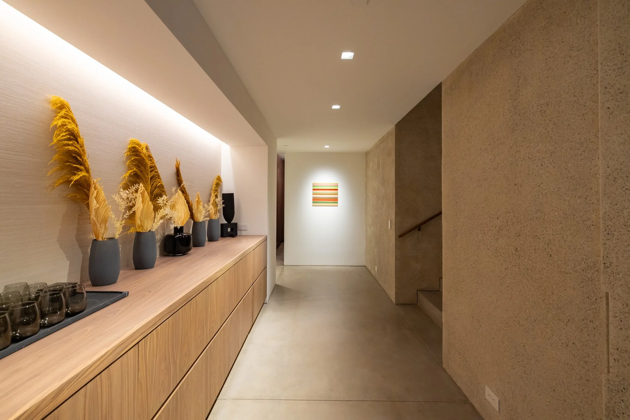 A modern indoor hallway with neutral-colored walls, a wooden sideboard decorated with vases and dried plants, and artwork hanging on the far wall under ceiling lights.
