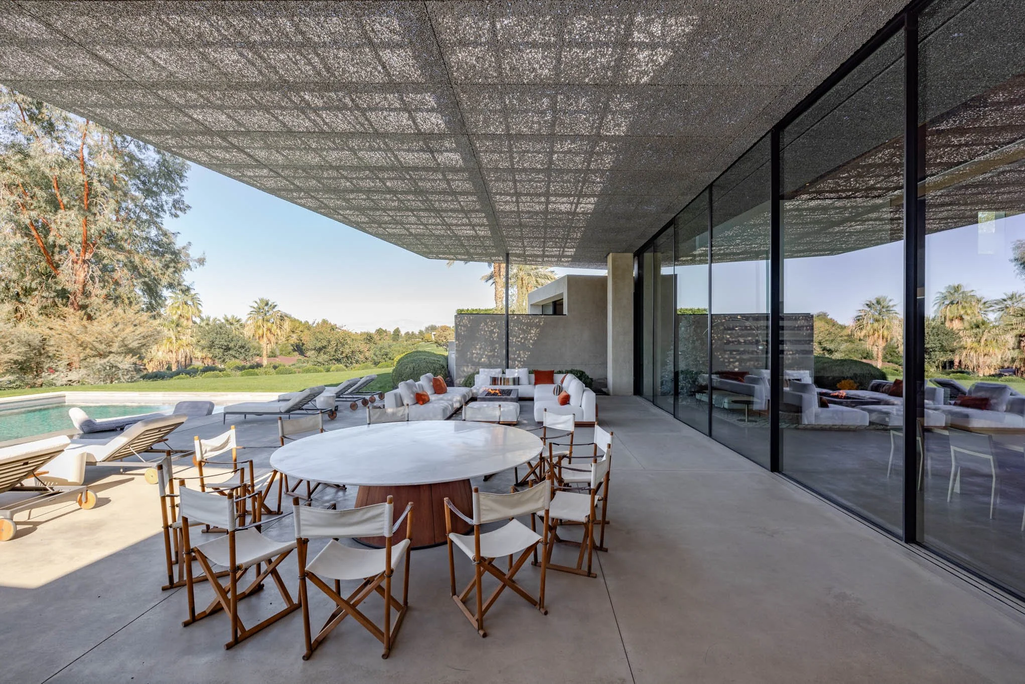 A modern outdoor patio with poolside lounge chairs, a round dining table with wooden chairs, white sofas with orange pillows, and large glass sliding doors reflecting the patio. The area overlooks a lush backyard with various trees and a swimming poo