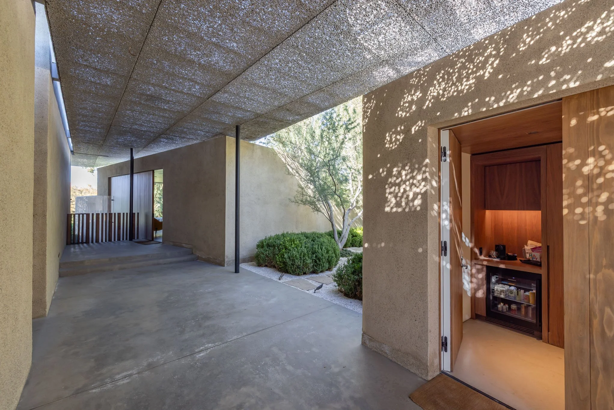 Covered outdoor area with concrete flooring, stucco walls, and a wooden door. Small landscaped garden with bushes and a tree. Dappled sunlight creates patterns on the wall.