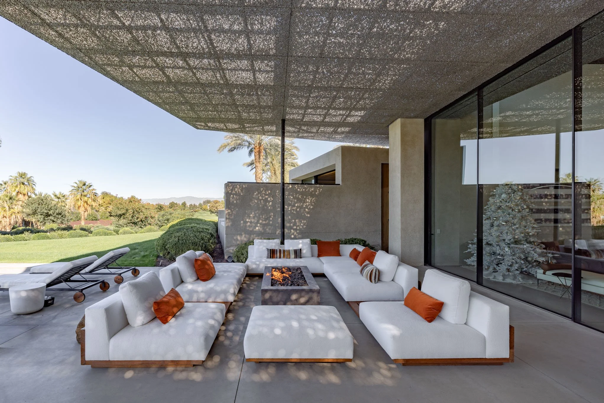 Modern outdoor patio with white sectional sofas, orange and striped throw pillows around a fire pit, black lounge chairs, and a view of green grass, shrubbery, palm trees, and distant mountains under a shaded ceiling.