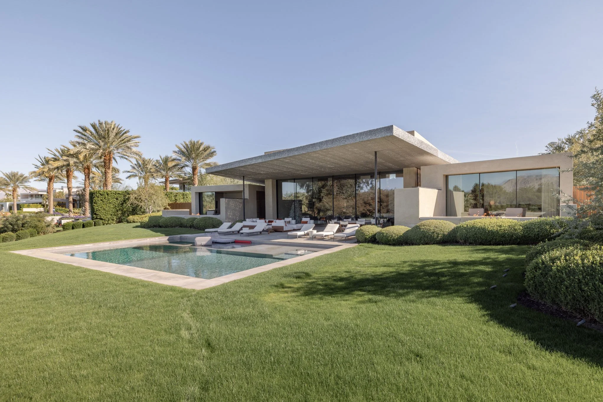 Modern house with large glass windows, covered patio, and a swimming pool surrounded by a landscaped lawn with palm trees and bushes.