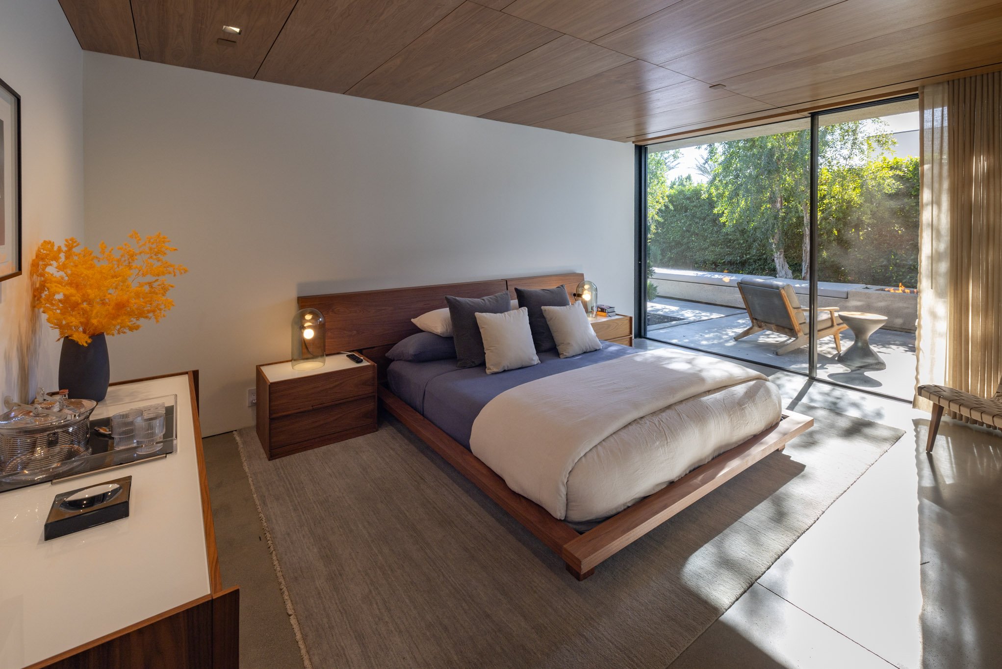 A modern bedroom with a large bed, gray and beige pillows, bedside tables with lamps, a sliding glass door leading to an outdoor patio with a chair and table, and a wooden ceiling.