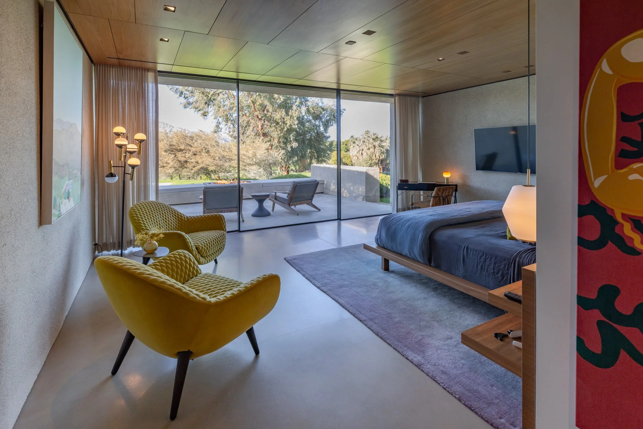 Modern bedroom with a large glass sliding door leading to an outdoor patio, featuring a bed with a dark duvet, a wall-mounted TV, a side table with a lamp, two yellow lounge chairs, a rug, and outdoor seating with a view of trees and mountains.