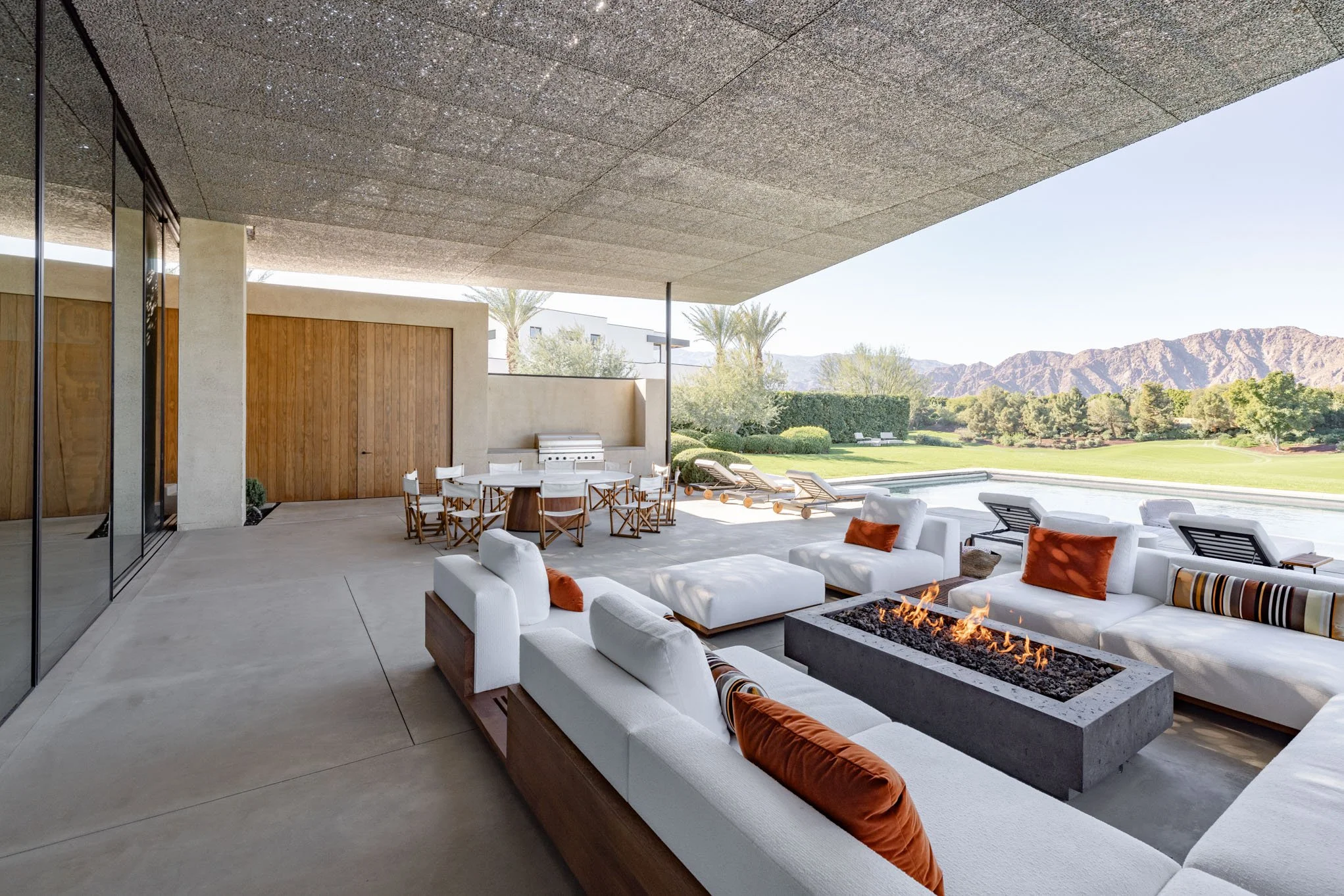 Modern outdoor patio area with white cushioned couches, a fire pit, a round dining table with chairs, lounge chairs by a pool, and a golf course with mountains in the background.
