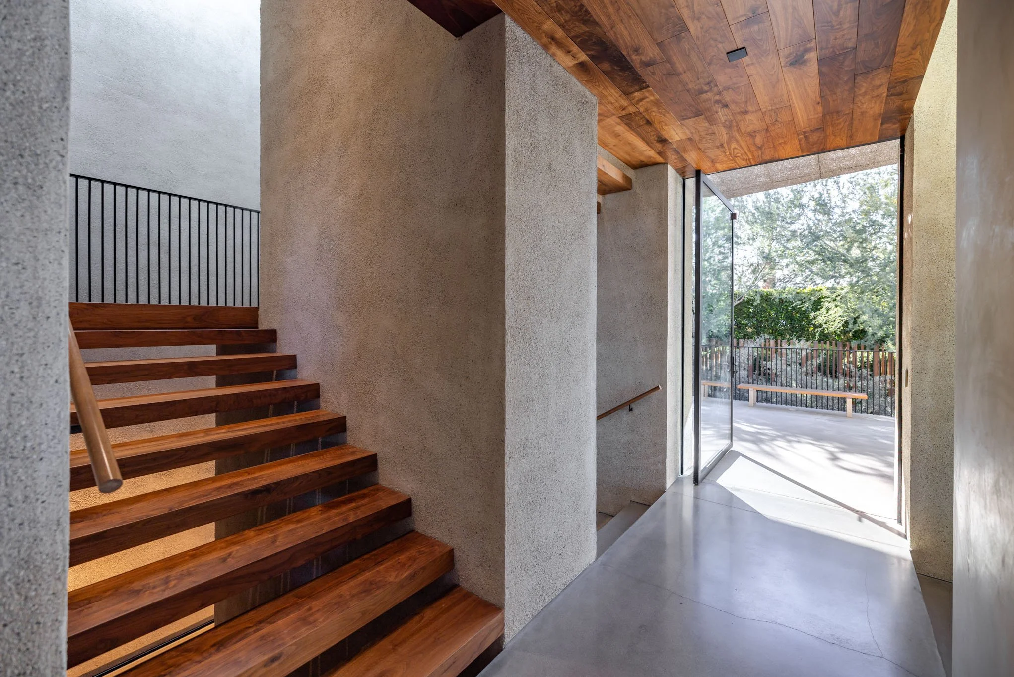 Modern interior hallway with wooden staircase and concrete walls leading to an exterior patio with trees and a wooden fence.