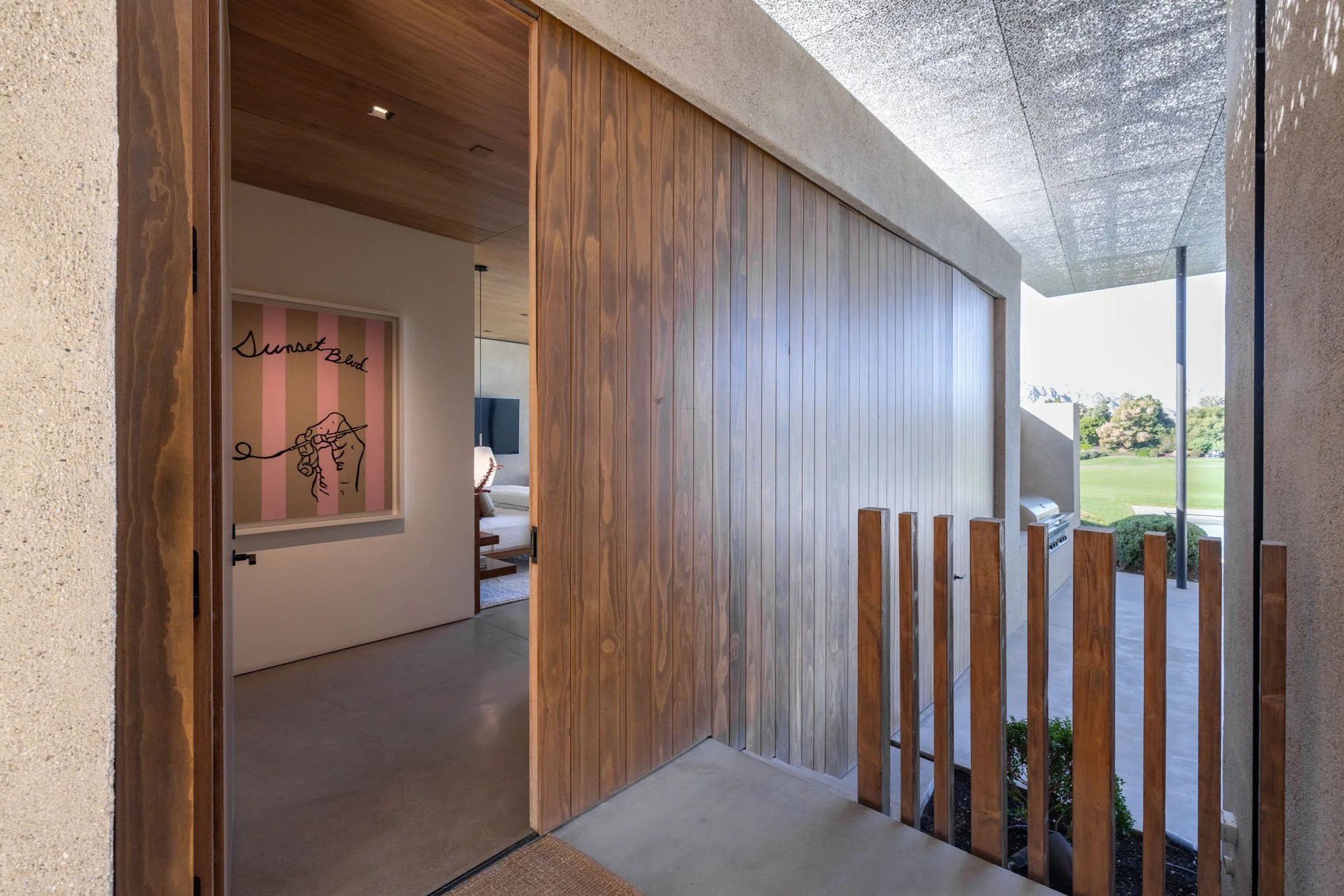 View from a covered porch into a modern house, showing interior through a doorway with a pink and white striped art piece and a bedroom with a bed, nightstand, and television.