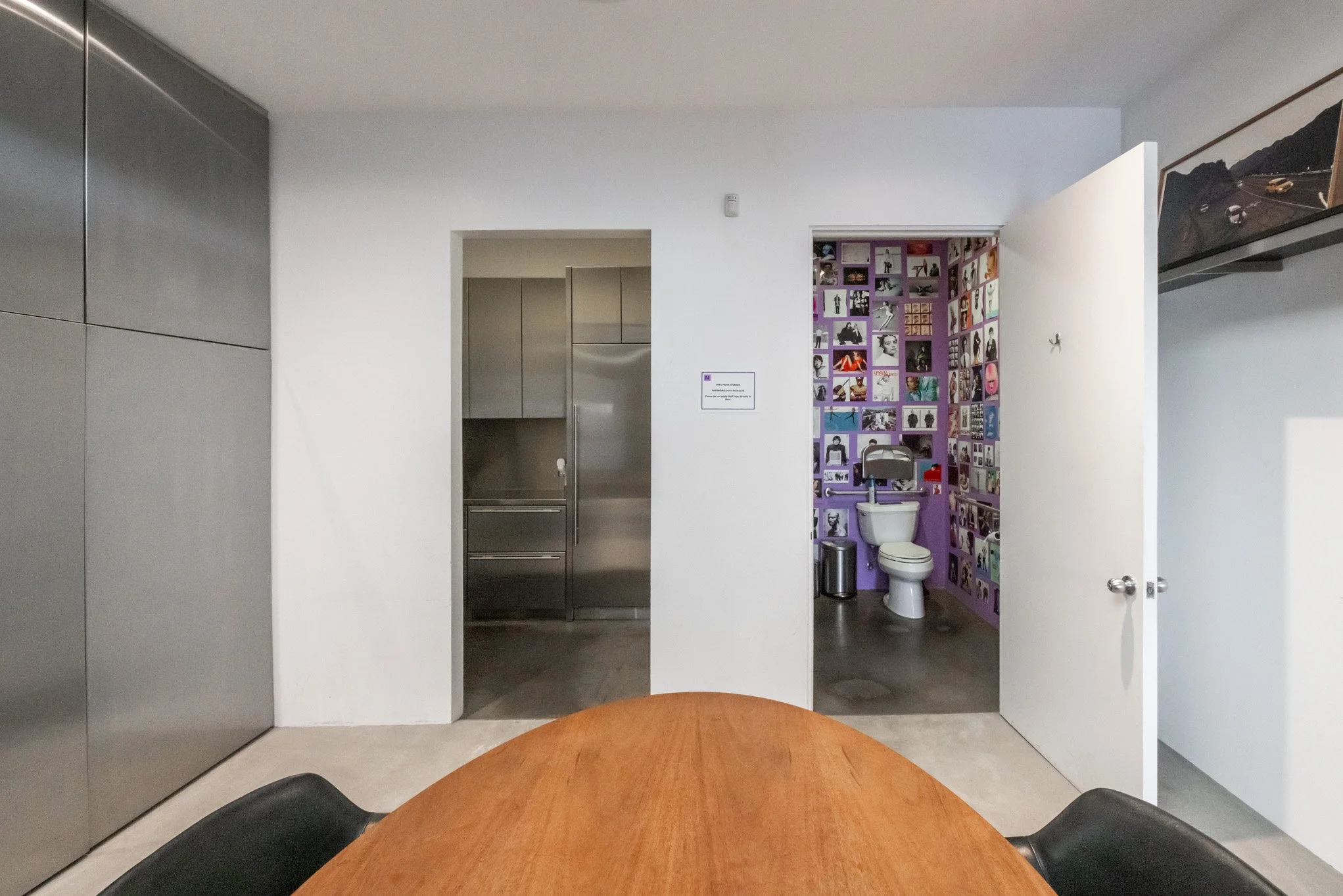 A modern interior room with a wooden table and black chairs, with a doorway leading to a bathroom with purple walls decorated with photos, a toilet, a metal trash can, and a small wastebasket.