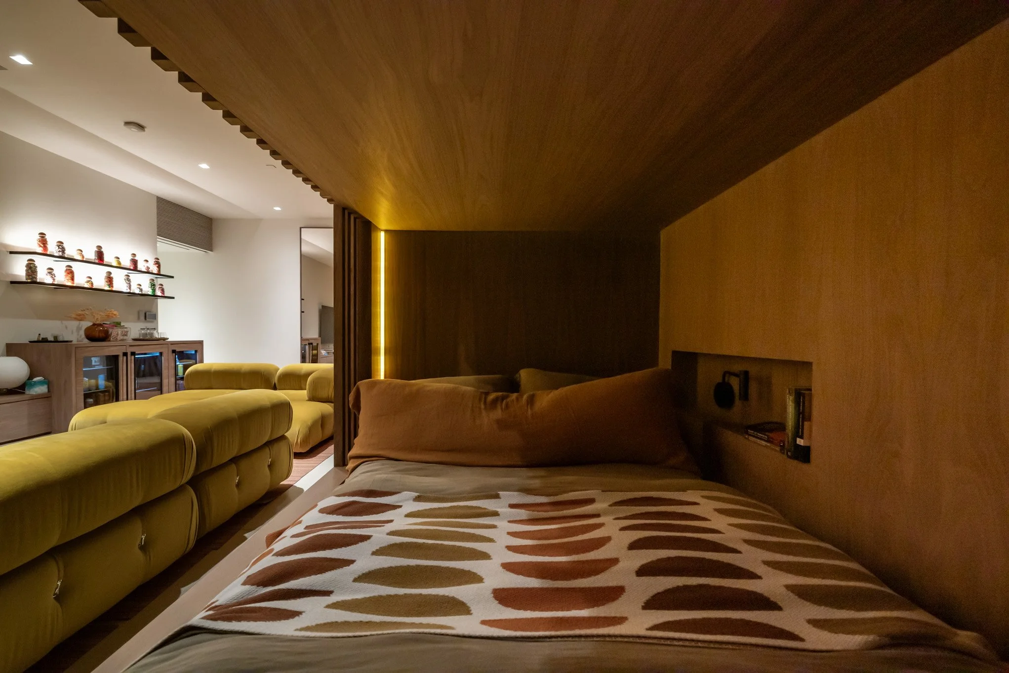 A cozy bedroom with wooden walls and ceiling, featuring a bed with a patterned blanket and blankets, a small built-in shelf with books, and a soft, warm lighting strip. In the background, a living area with a yellow sofa and a white wall with shelves