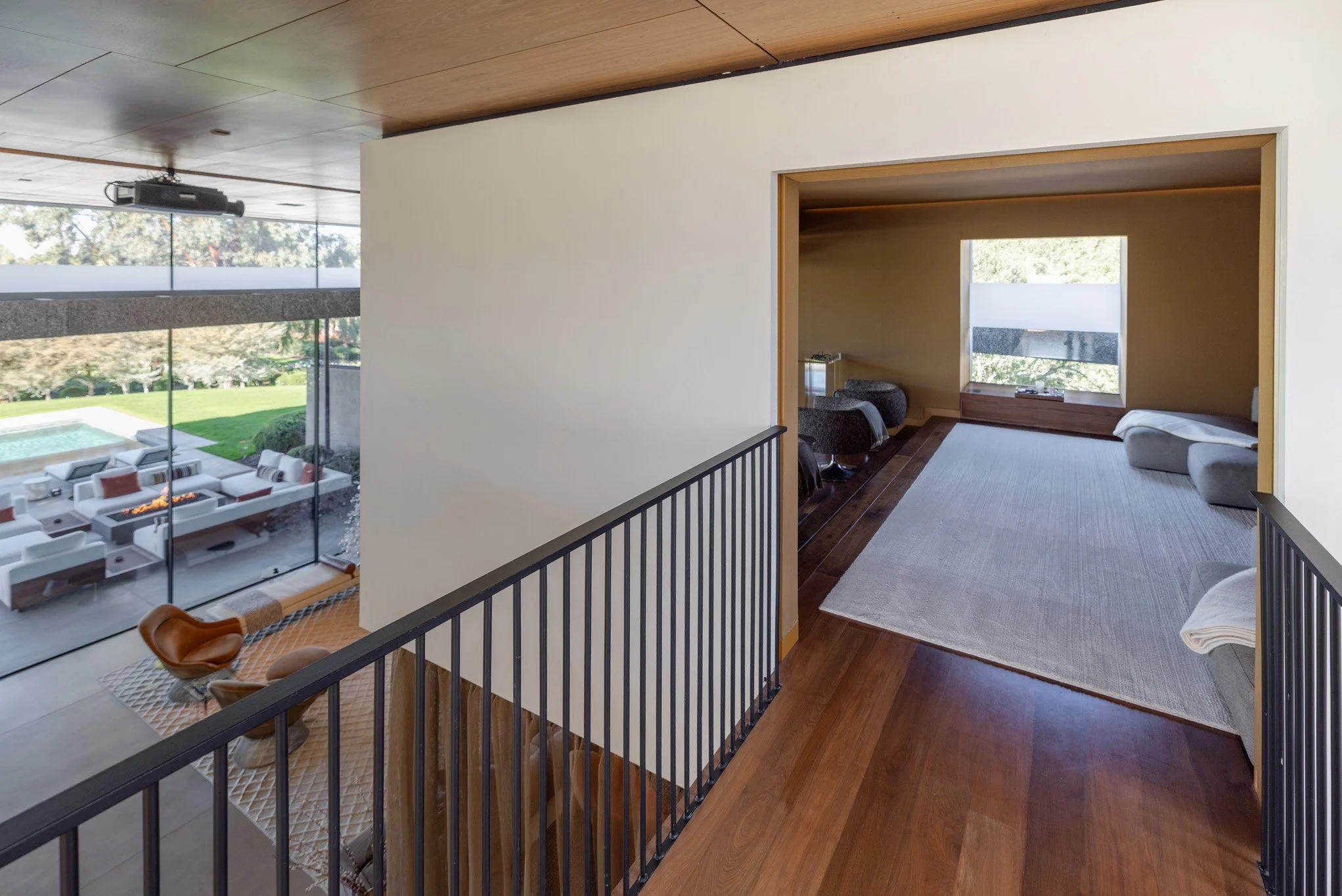 Interior view of a modern house with a hallway overlooking living space with large windows, an outdoor patio, seating, and greenery outside.