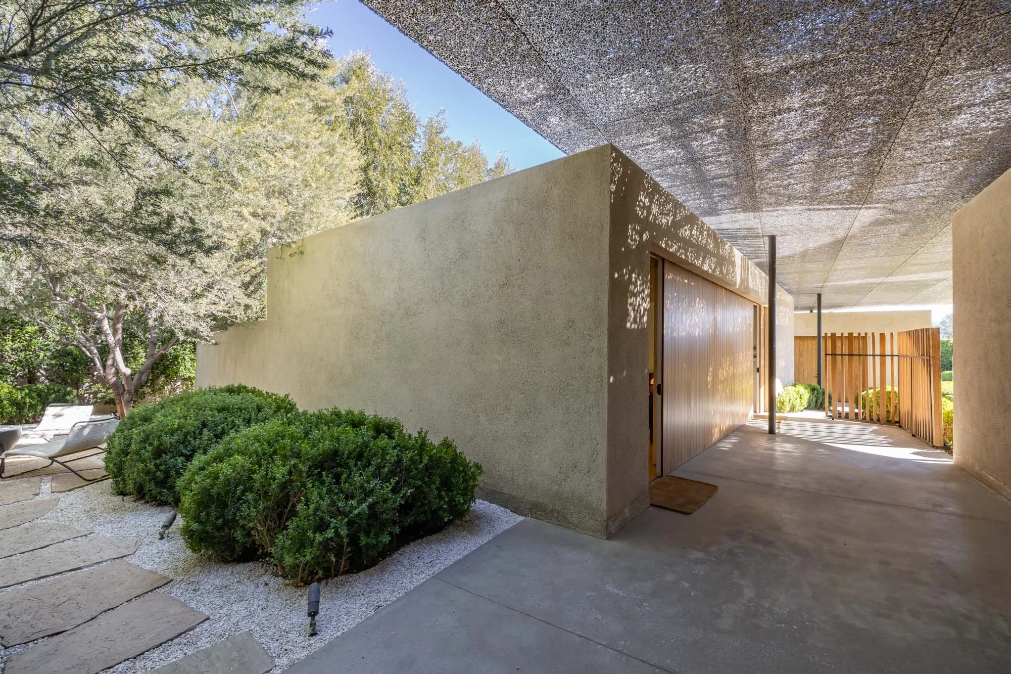 Modern house exterior with a covered outdoor corridor, concrete pathway, greenery, and wooden fence