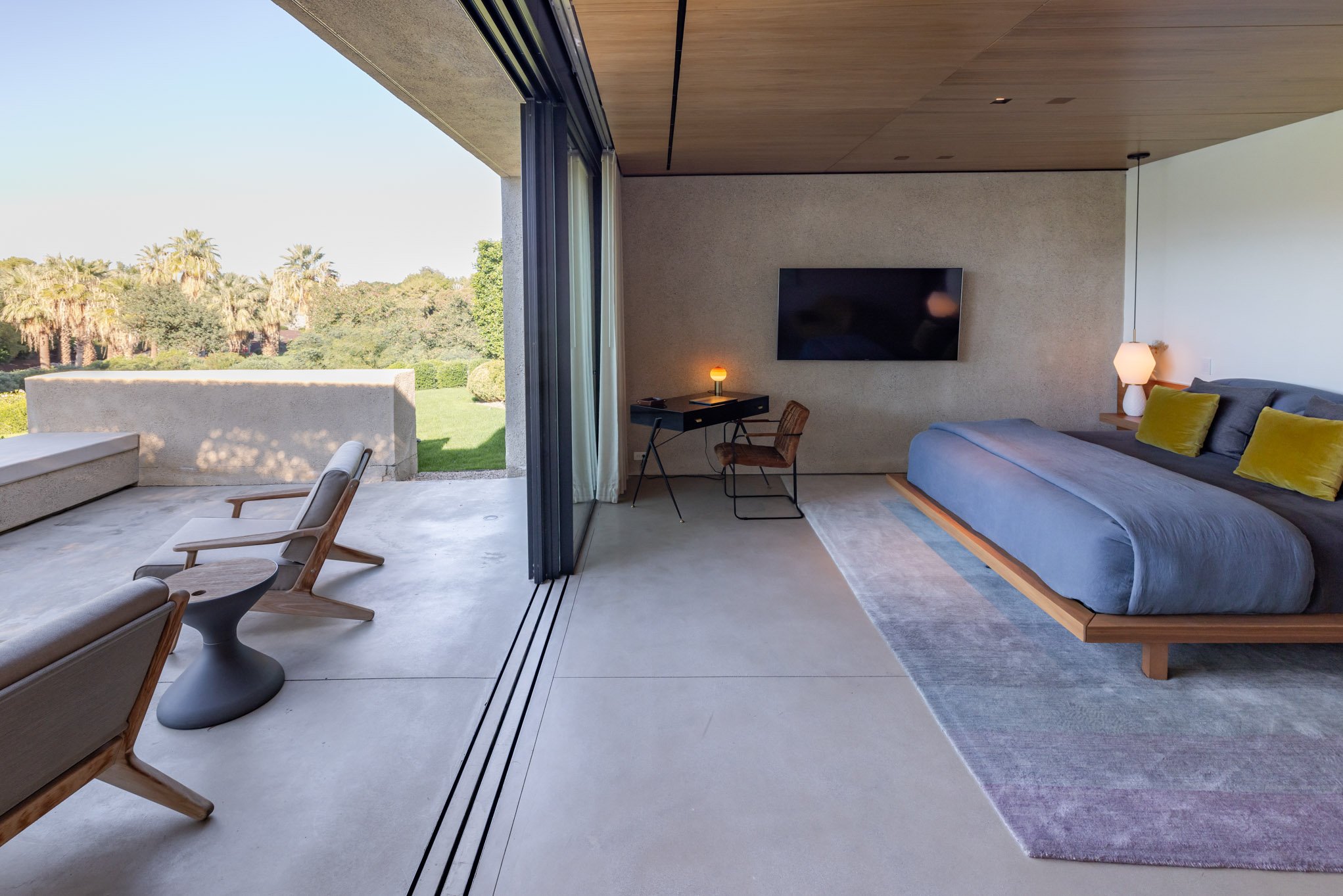 Modern bedroom with large glass doors opening to a patio and green garden. Inside, a bed with gray and yellow pillows, a small side table with a lamp, a flat-screen TV on the wall, a small desk with a lamp, and a chair. Outside, patio furniture with 