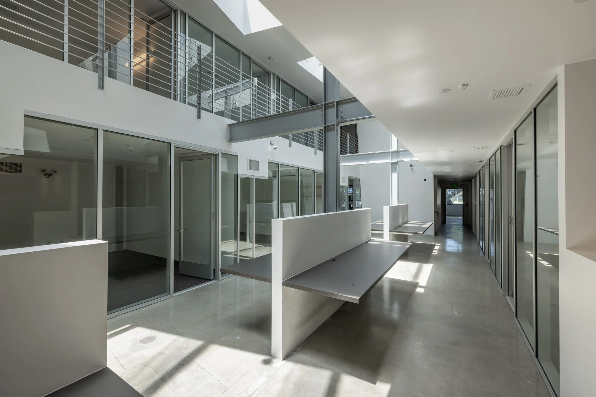 Modern, minimalist interior hallway with glass walls, metal railings, and benches, illuminated by natural light.
