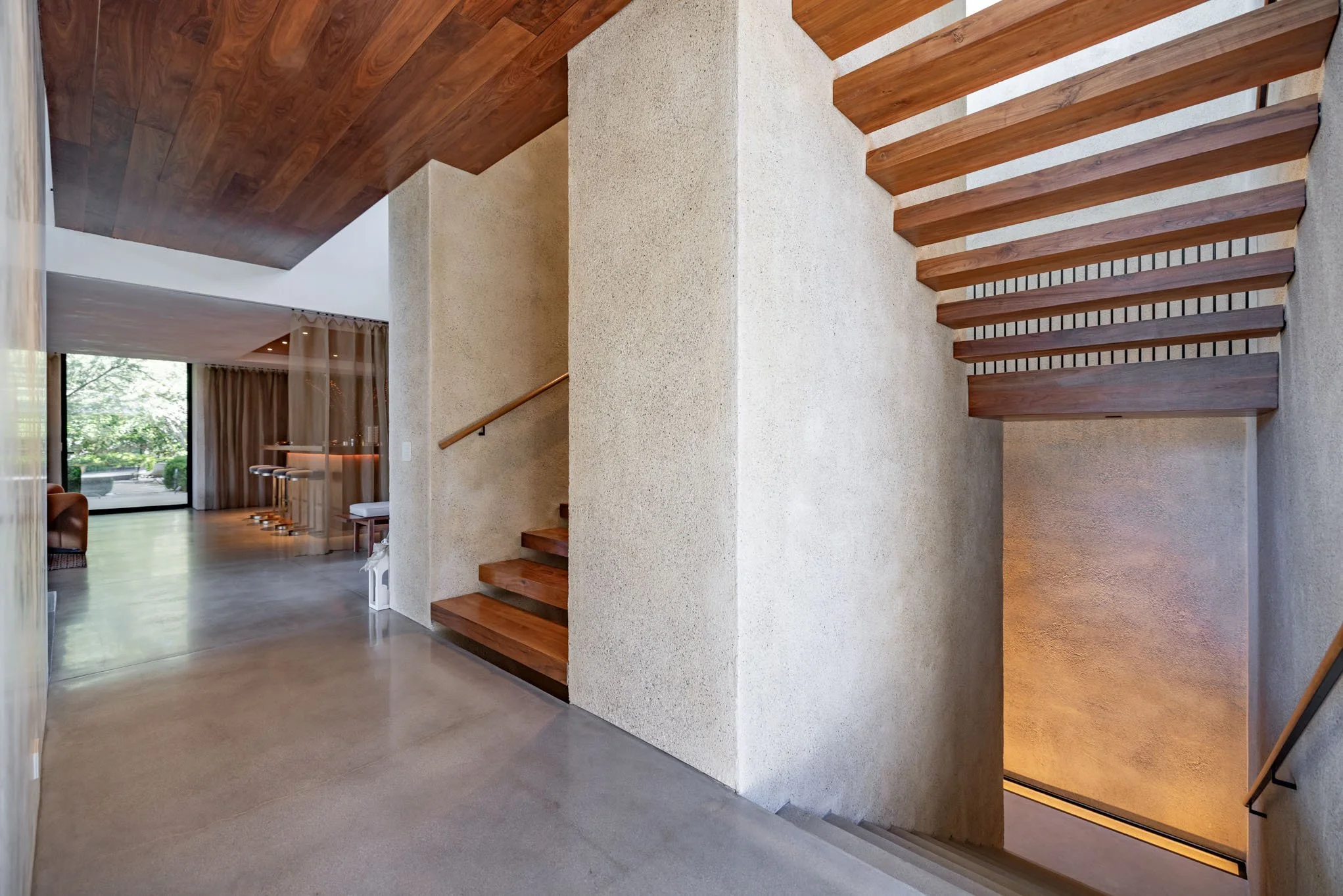 Interior view of a modern home featuring a staircase with wooden steps and a textured wall, adjacent to a living area with large windows and curtains.
