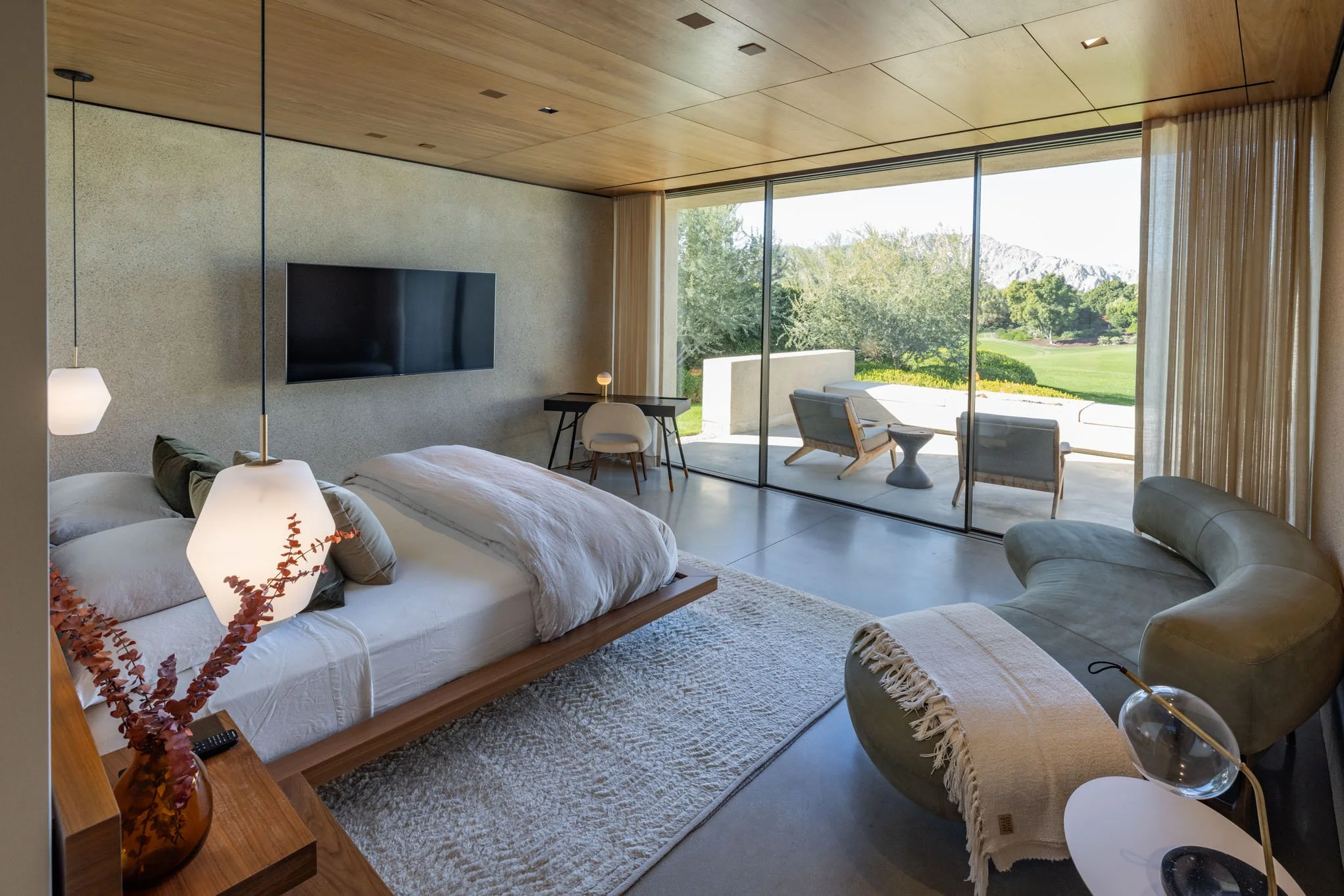 Modern bedroom with large sliding glass doors leading to an outdoor patio with seating and view of a green landscape and mountains.