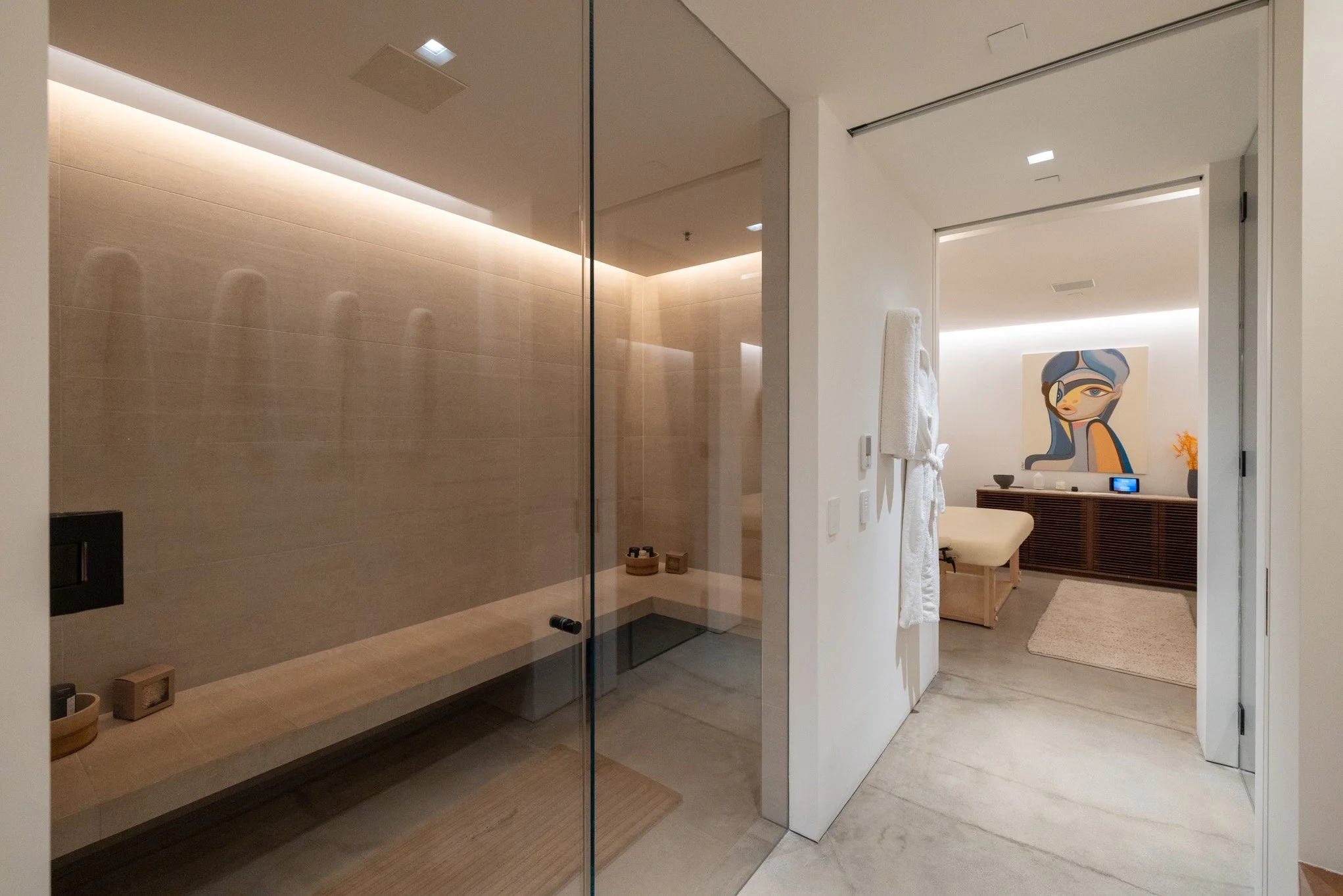 Inside a modern, minimalist bathroom with a glass shower and a hallway leading to a room with a colorful abstract painting and a desk.