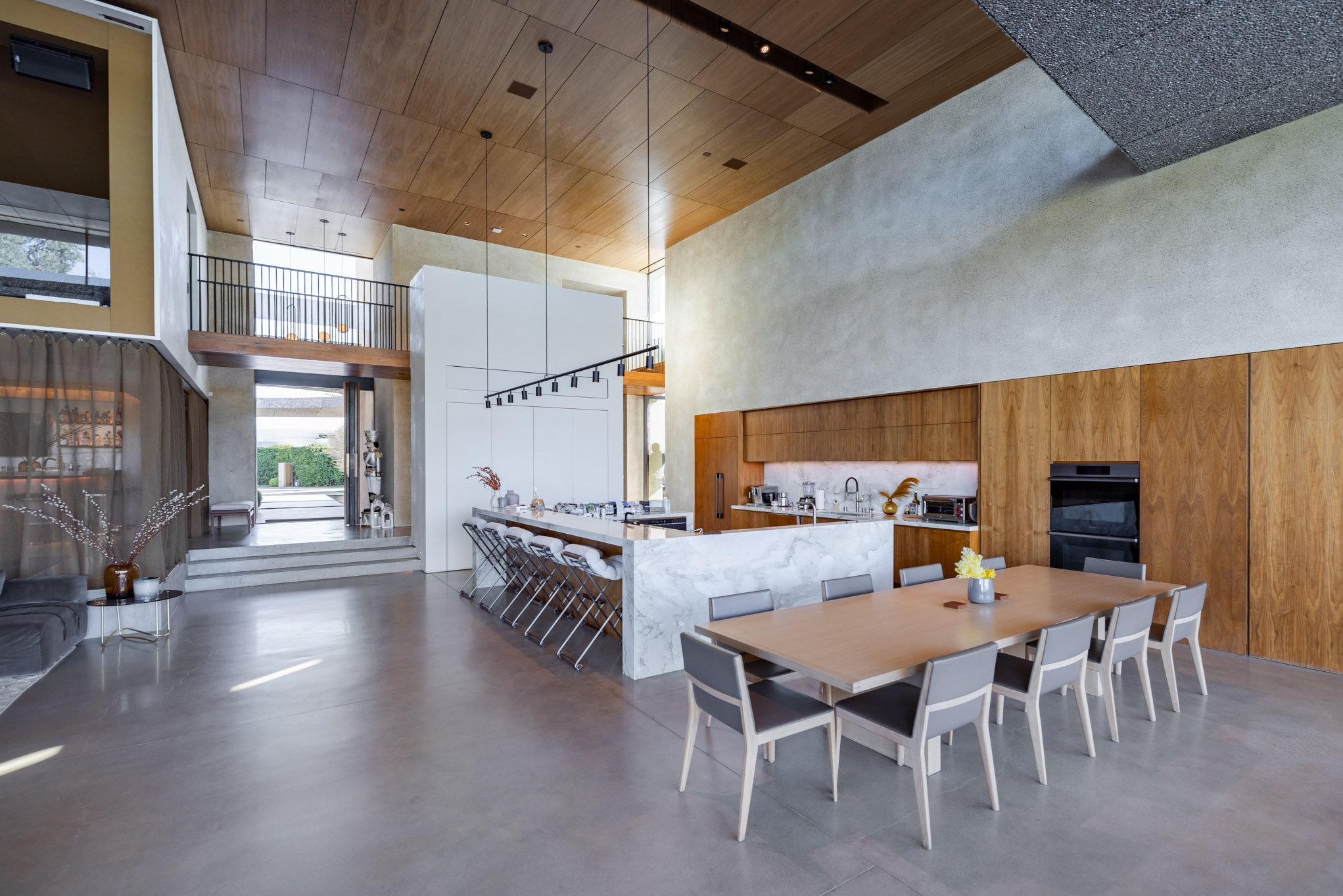 Open-concept modern kitchen and dining area with wooden accents, marble countertops, and a high ceiling.