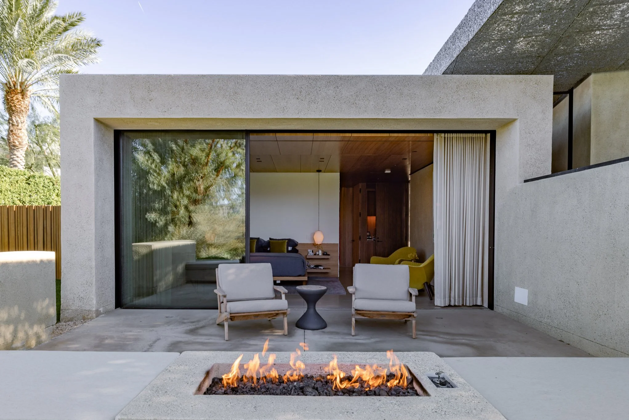 Modern house with glass sliding doors, outdoor seating with two white chairs, fire pit in the foreground, and view into a stylish living room with green armchair and bedroom with bed and pendant light