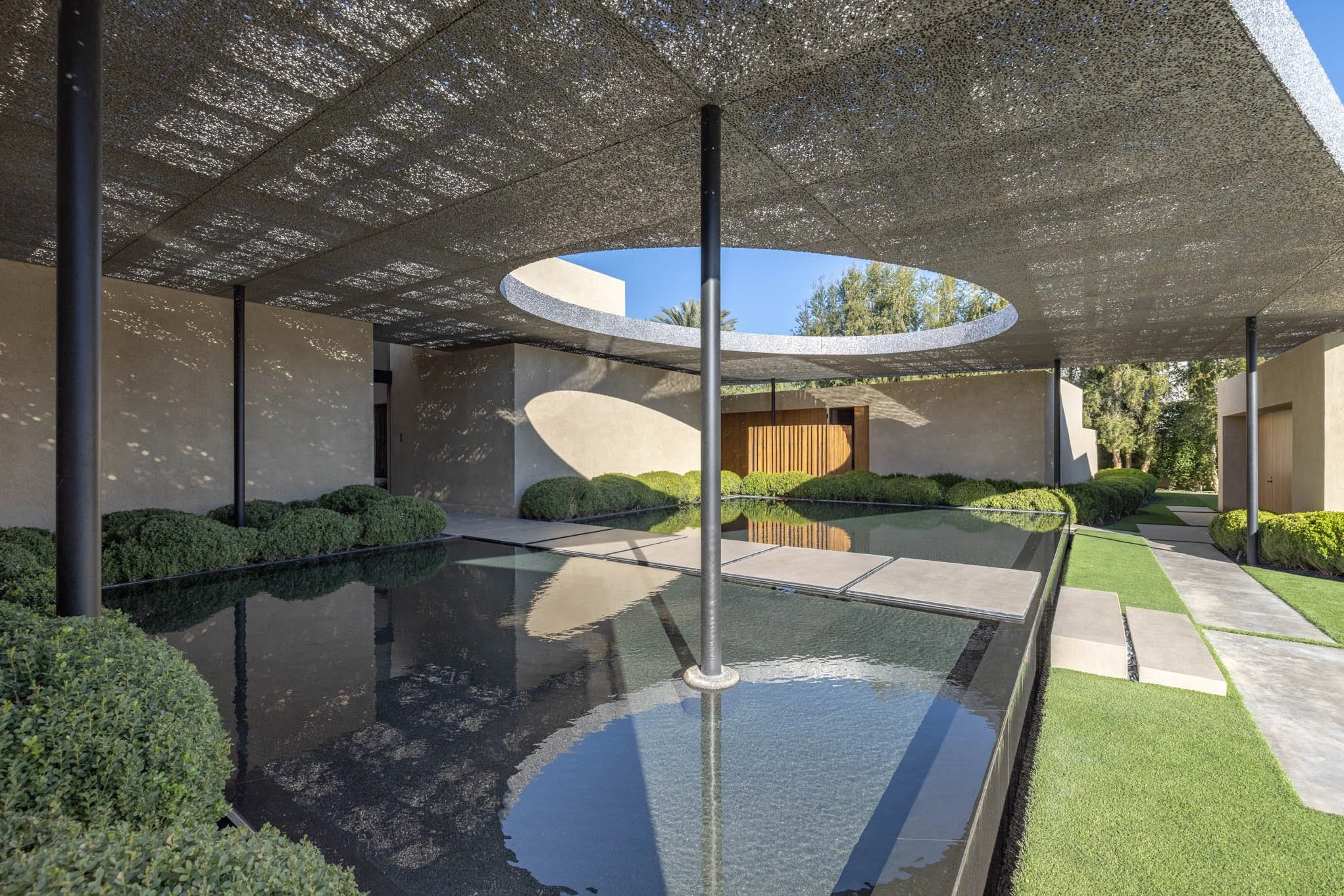Modern outdoor structure with a large circular opening in the ceiling, reflecting pool, greenery, and pathways.
