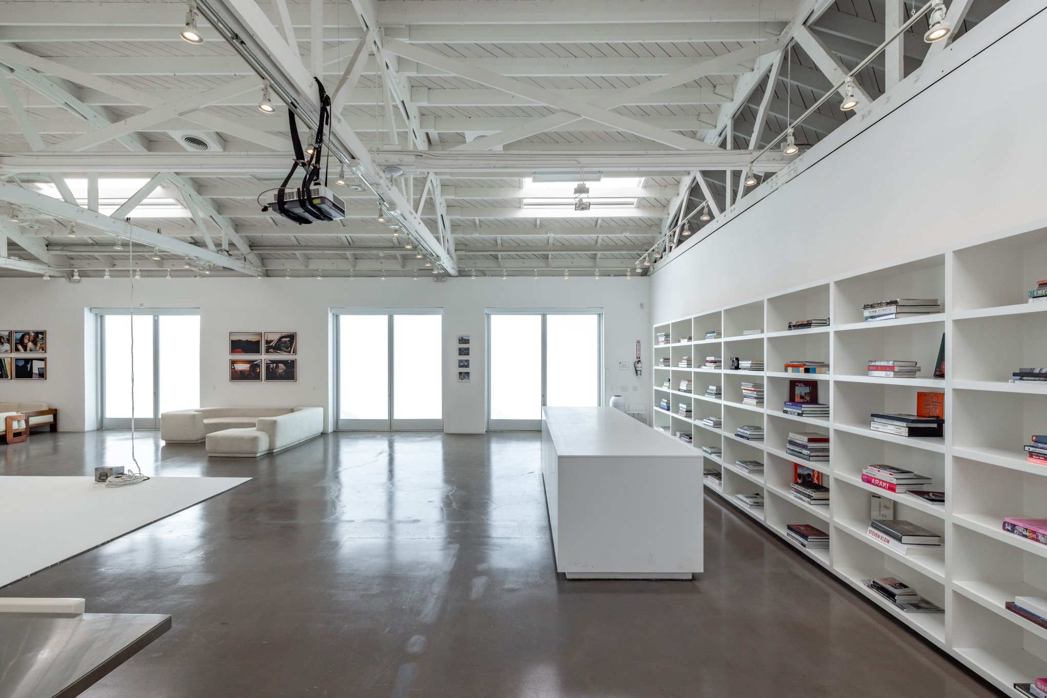 A spacious, modern room with white walls, ceiling, and shelves, featuring large windows and sliding doors allowing natural light, with a white couch and a wall-mounted TV, and a collection of magazines and books on open shelves.