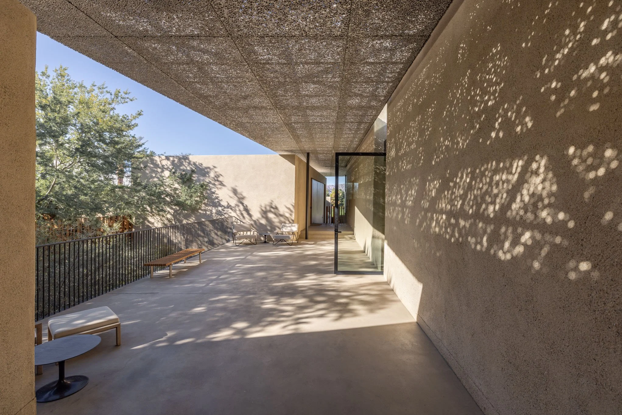 An outdoor patio with lounge chairs, a small table, and benches, shaded by trees with sunlight creating patterns on the wall and floor.